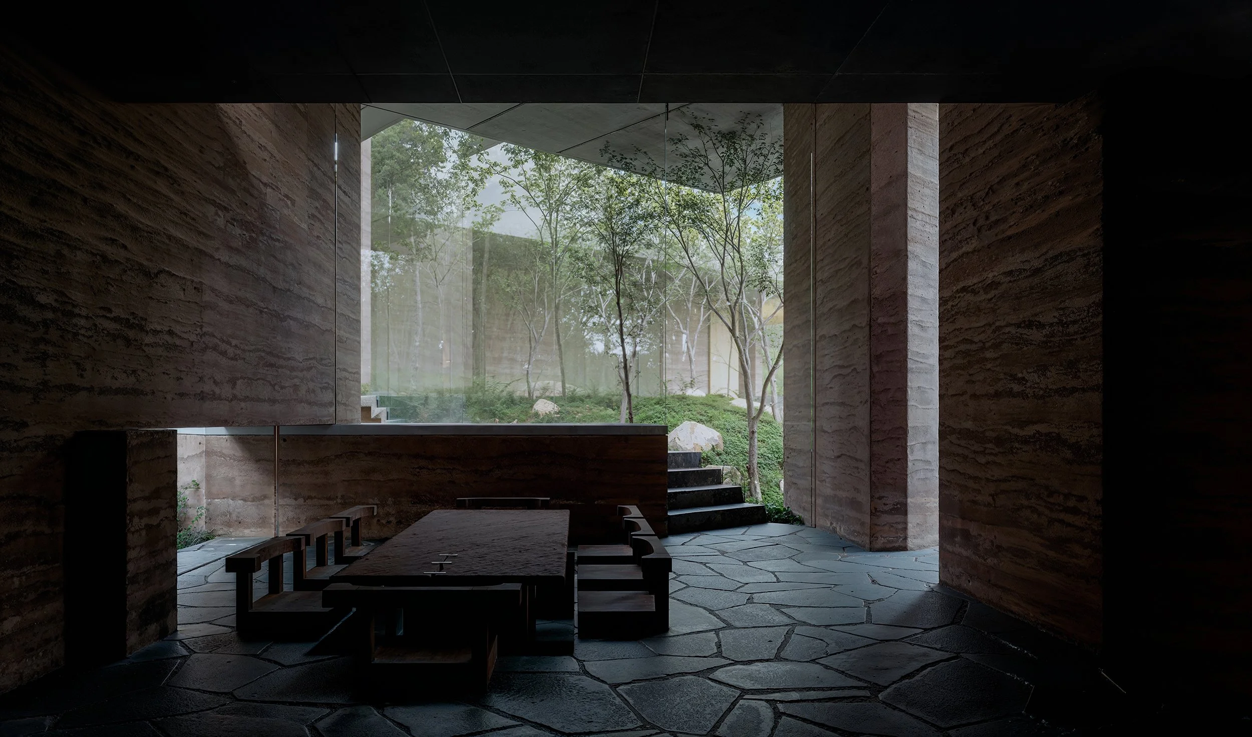 Interior view of a modern building with large glass window showing green trees outside. The room features stone walls, a stone floor with a geometric pattern, and wooden furniture including a table and chairs.