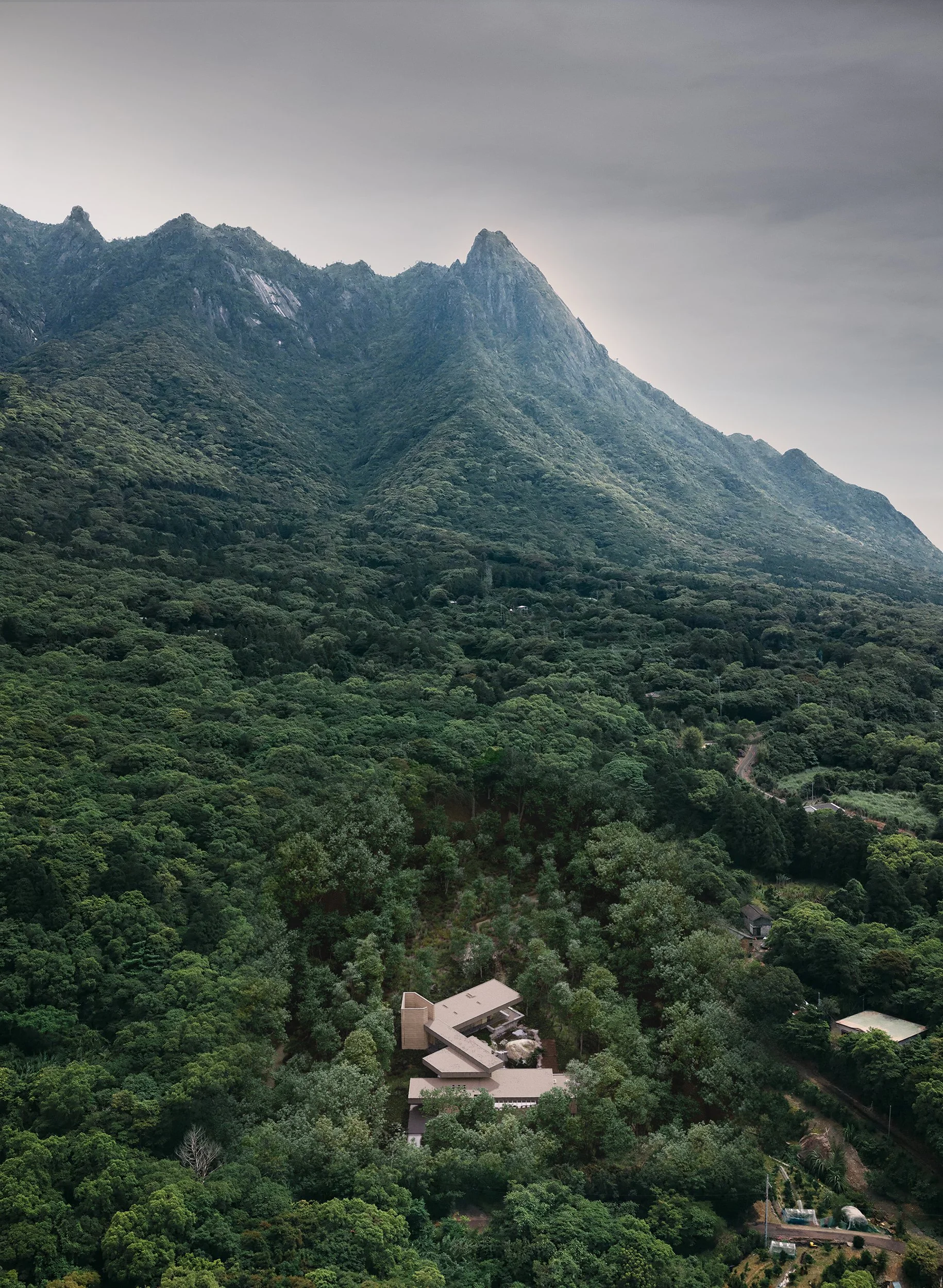 A mountain with dense green forest and a modern building complex at the mountain base.