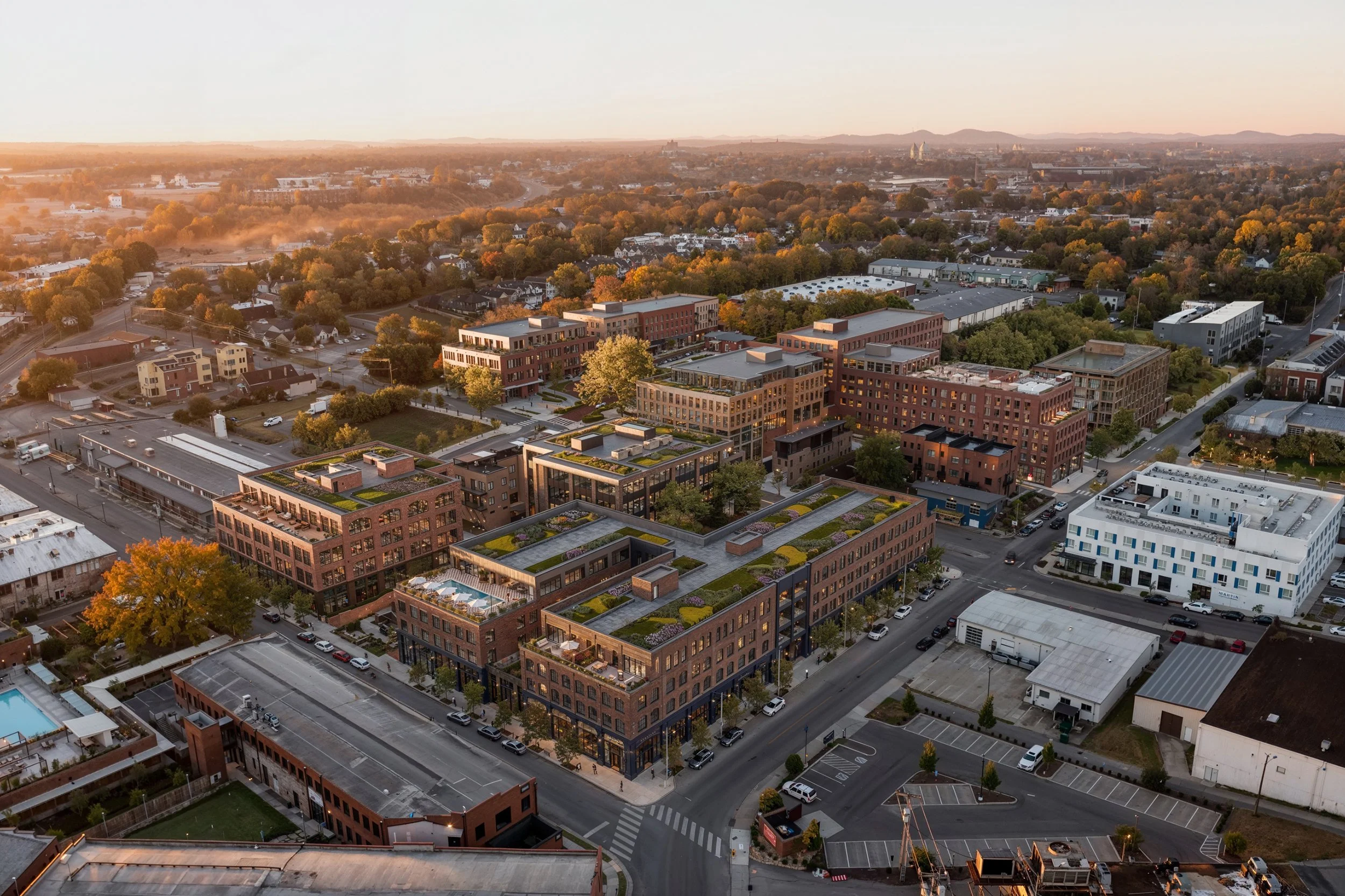 An aerial view of a cityscape during sunset with modern buildings, trees with fall foliage, and distant hills in the background.
