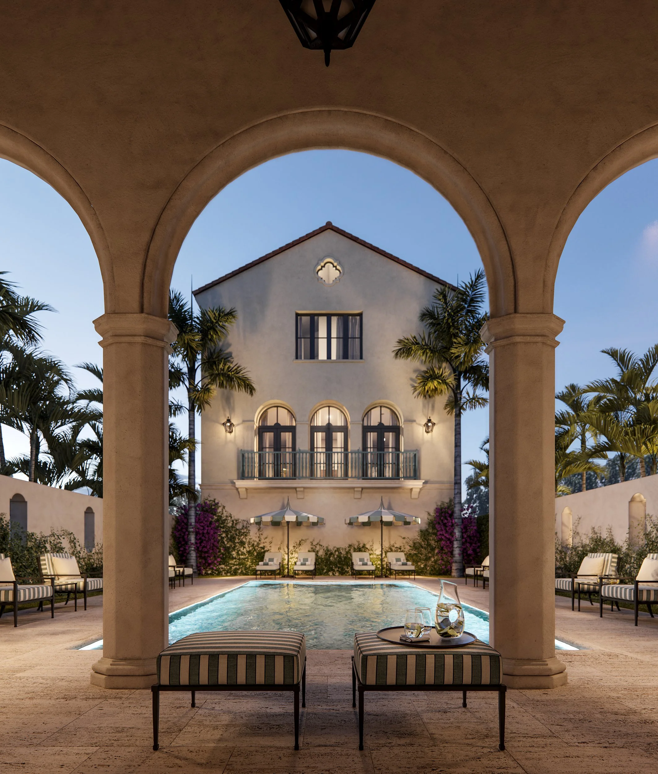 View of an outdoor swimming pool area at dusk, seen through large stone archways. There are lounge chairs and umbrellas around the pool, with a white building featuring arched windows and a balcony in the background. Palm trees border the scene, crea