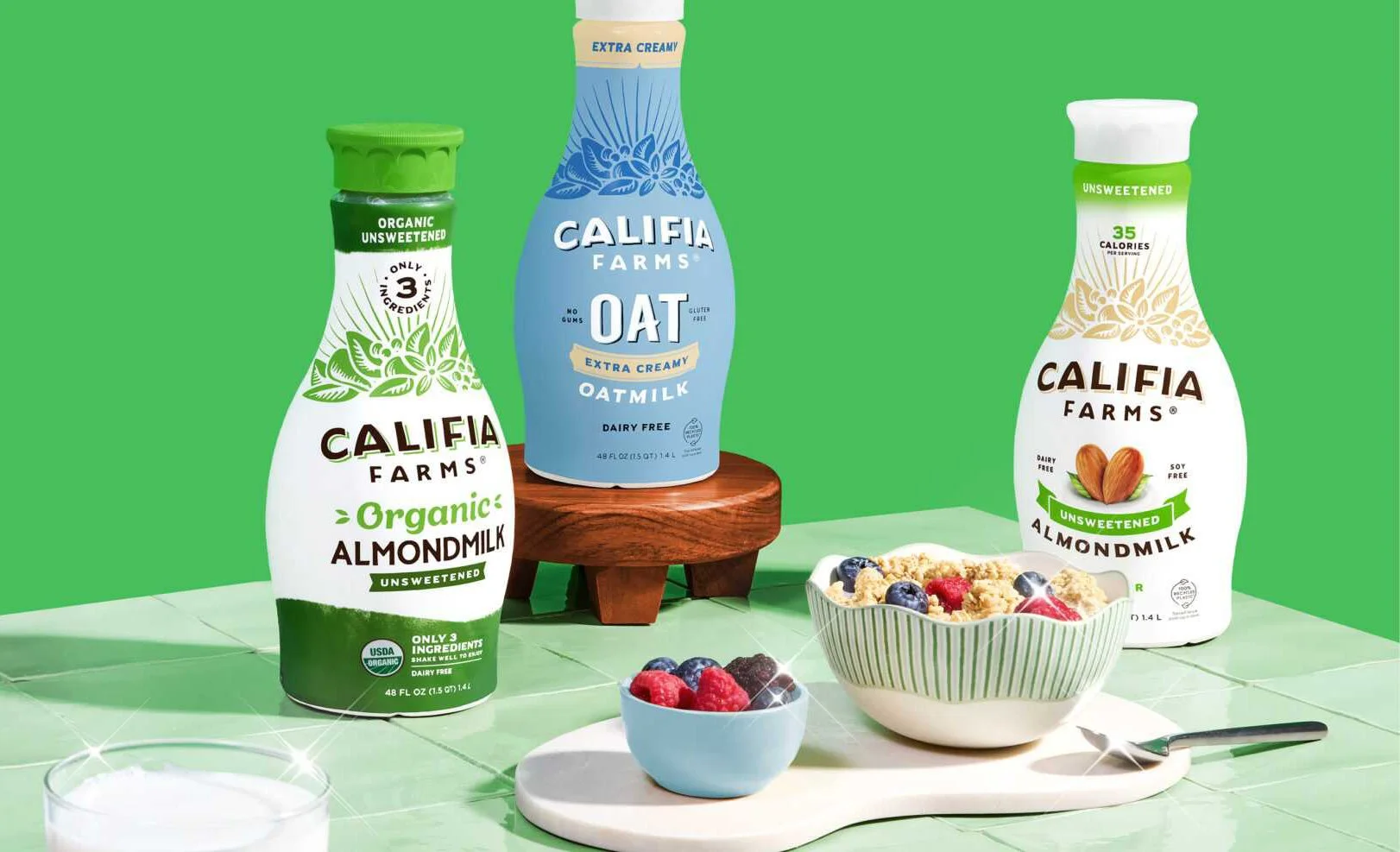 Three bottles of Califia Farms plant milk with different labels on a green background, alongside a bowl of mixed berries and a bowl of cereal, on a light green tiled surface.