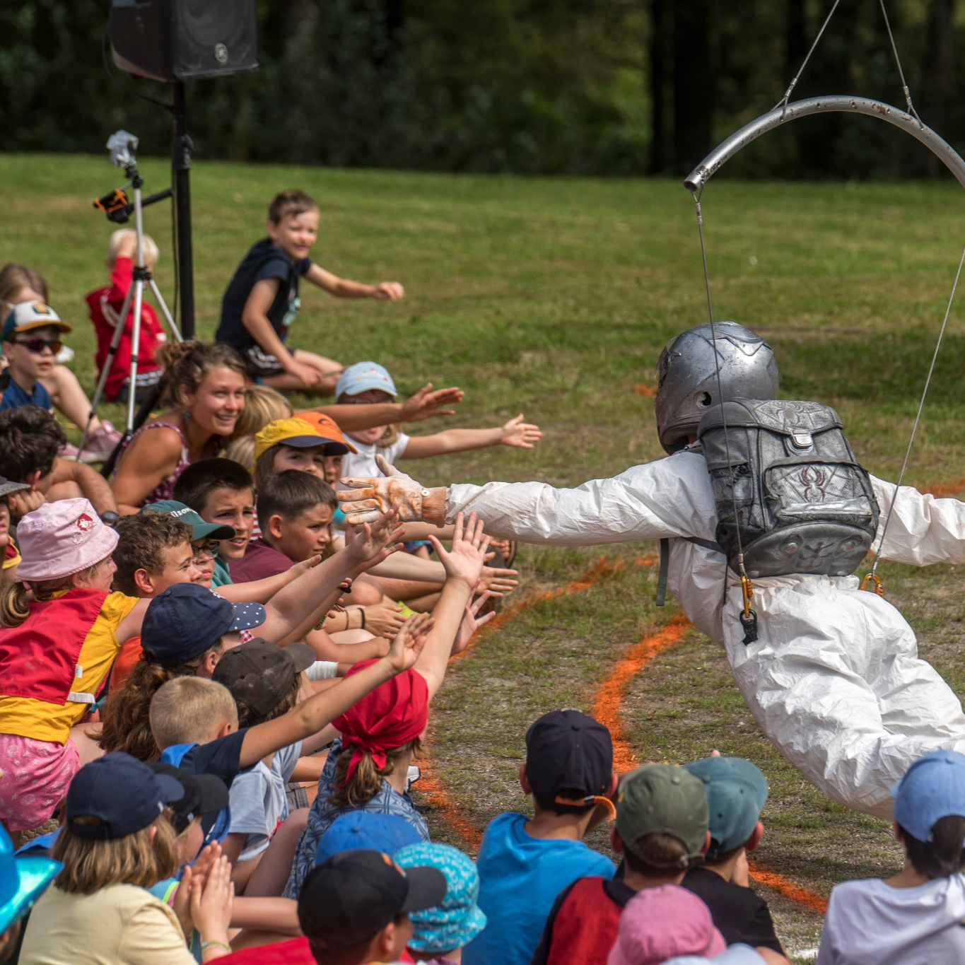 A person in a white protective suit, helmet, and backpack reaching out to children seated on the grass outdoors, with children extending their hands to shake his.