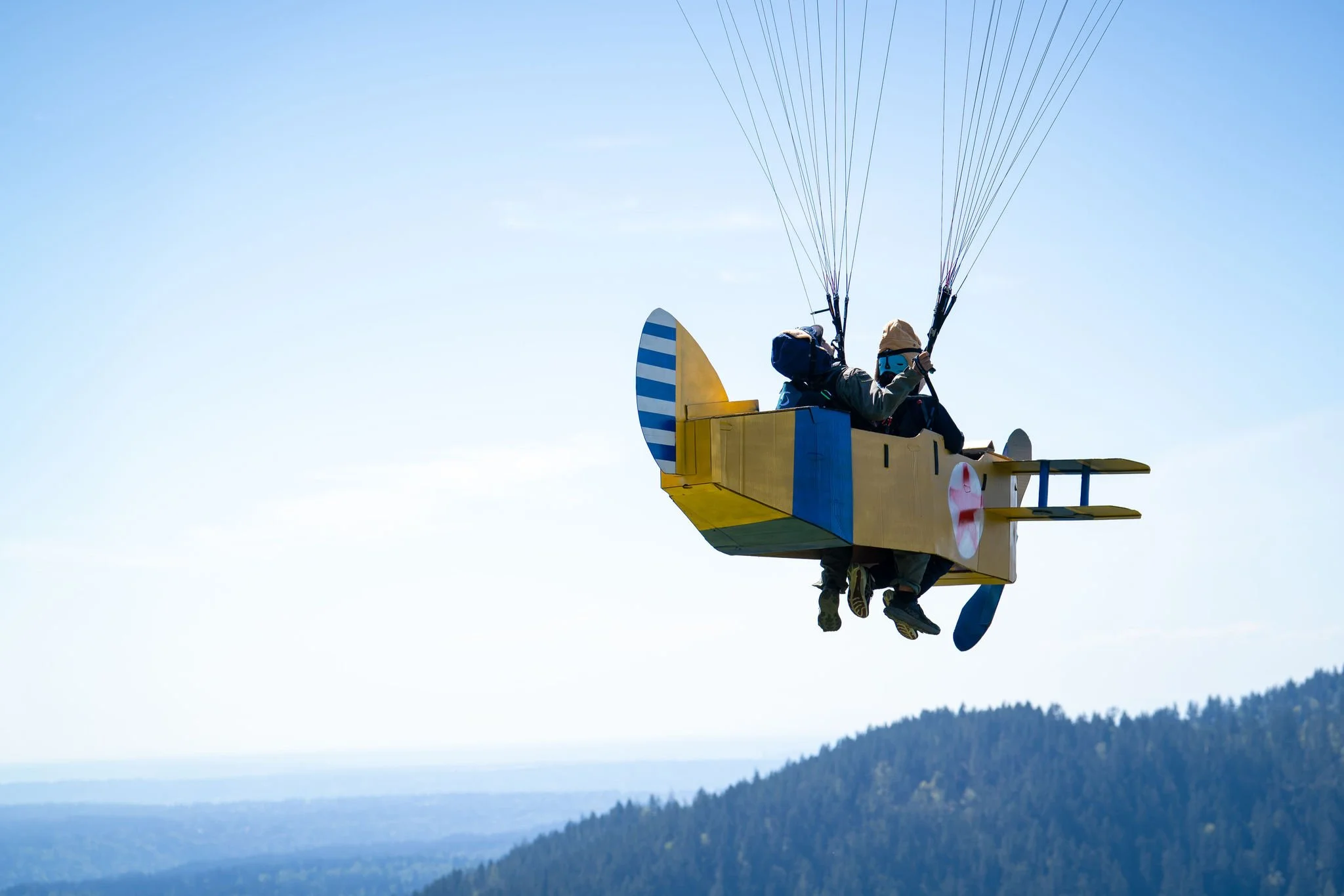 Two people riding a yellow powered hang glider or ultralight aircraft with blue and white striped wings over a mountainous landscape.