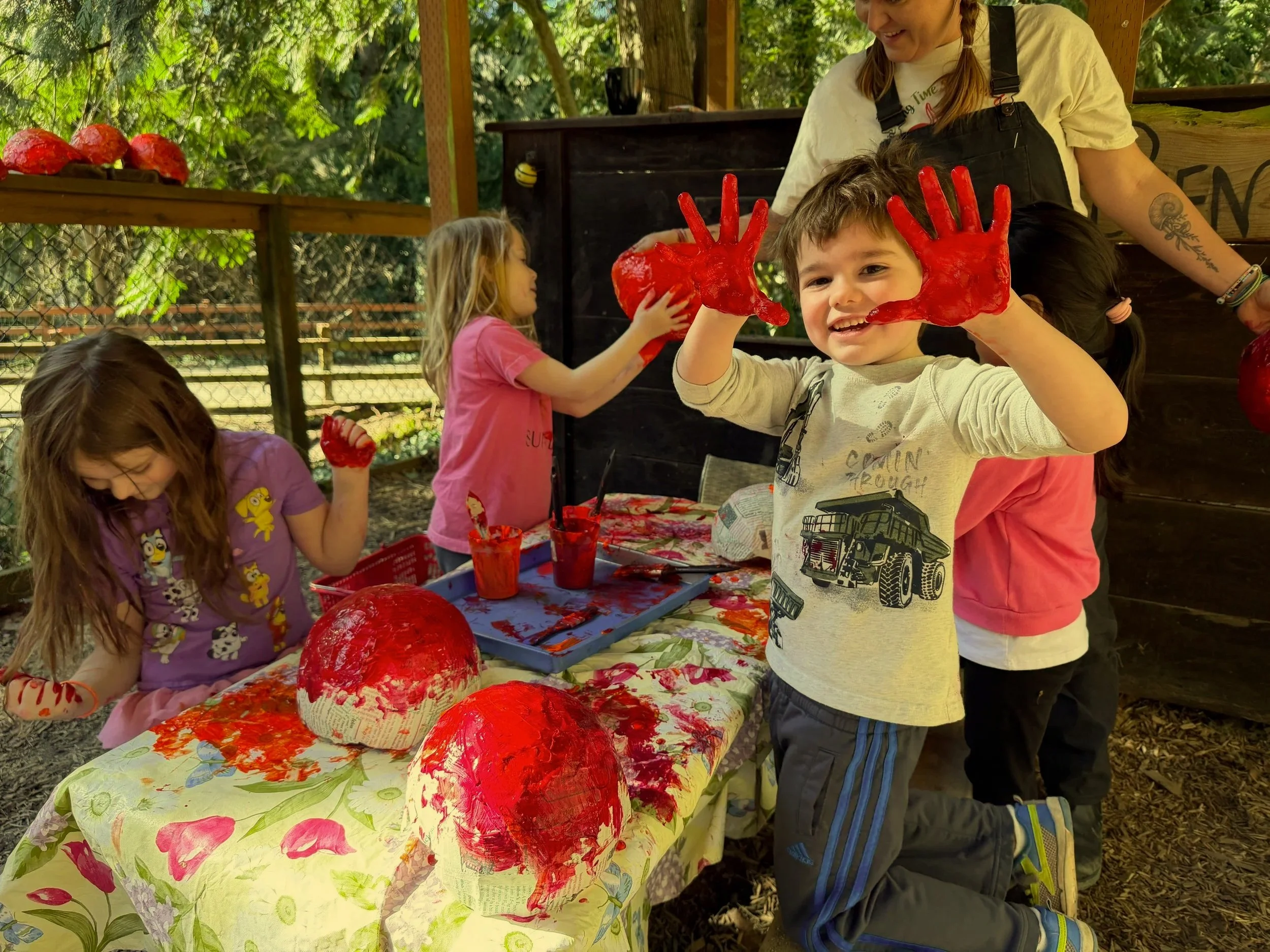 Children at an outdoor craft table with red painted papier-mâché pumpkins, supervised by an adult woman, surrounded by trees and a fenced yard.