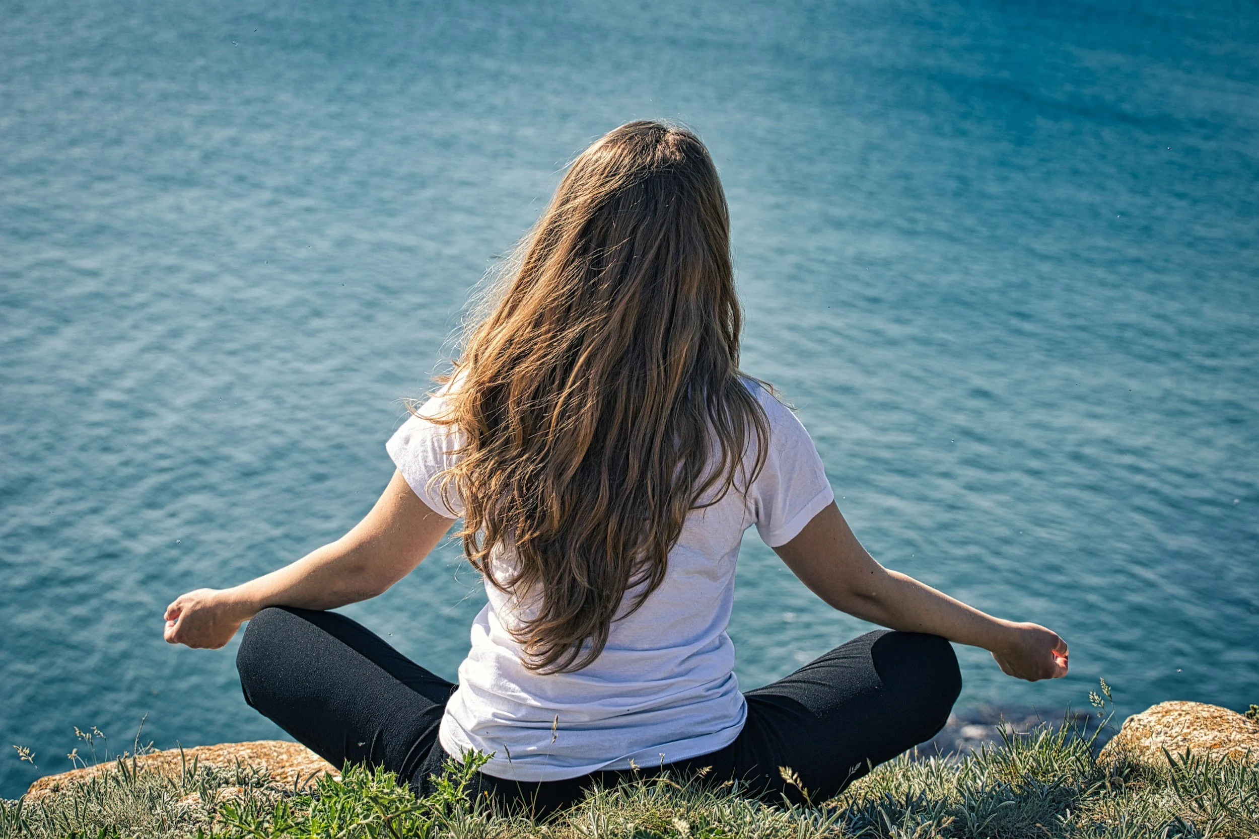 A woman with long wavy hair practicing yoga outdoors near water, sitting cross-legged on grass and facing the water.