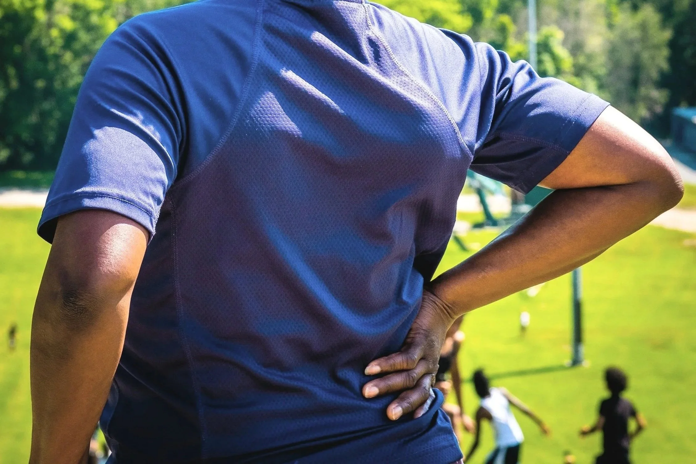 Close-up of a person wearing a blue athletic shirt with their hand on their hip, outdoors in a green park with trees and a playground in the background.