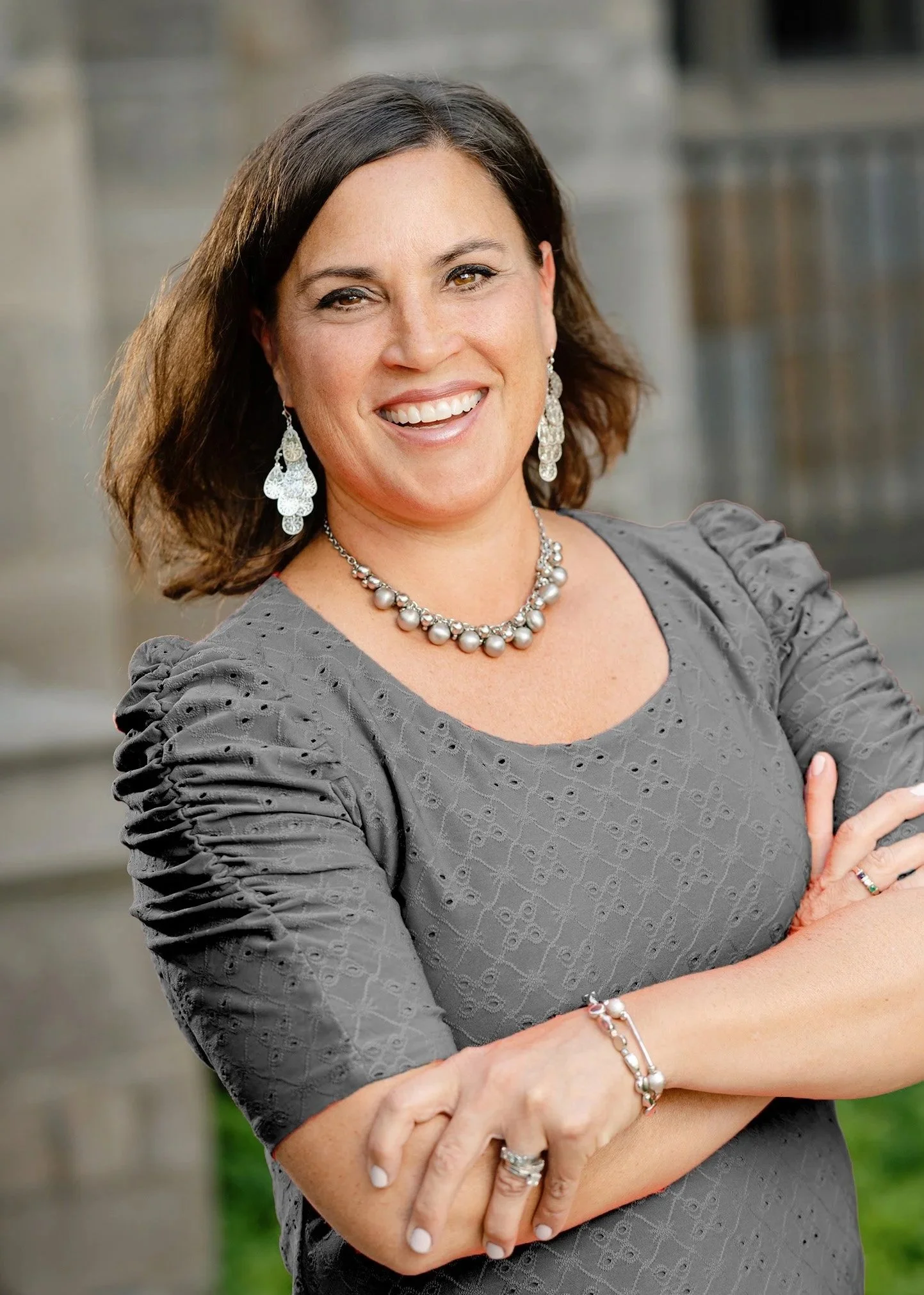 A woman with dark brown hair and light skin smiling, wearing a gray dress with ruched details, large pearl and silver jewelry including earrings, necklace, bracelet, and rings, with crossed arms, outdoors with blurred background.