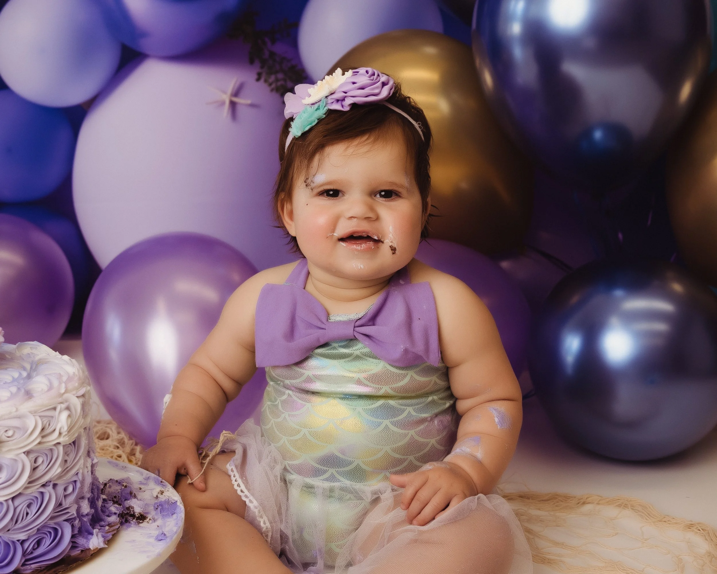 A young girl sitting beside a decorated birthday cake with purple and white icing, surrounded by purple and gold balloons, at a birthday celebration.