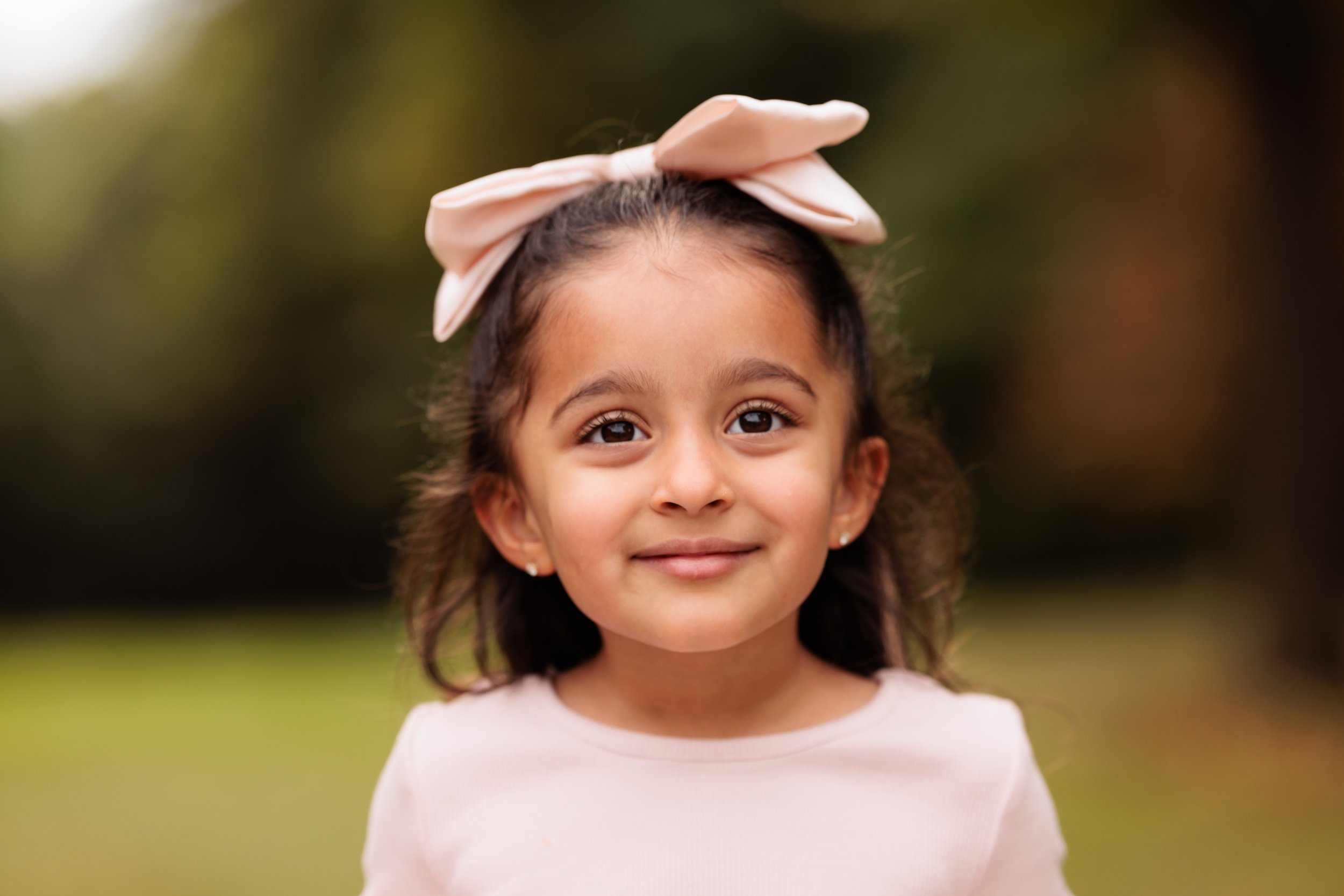 Young girl with a large pink bow on her head, smiling outdoors with blurred greenery in the background.
