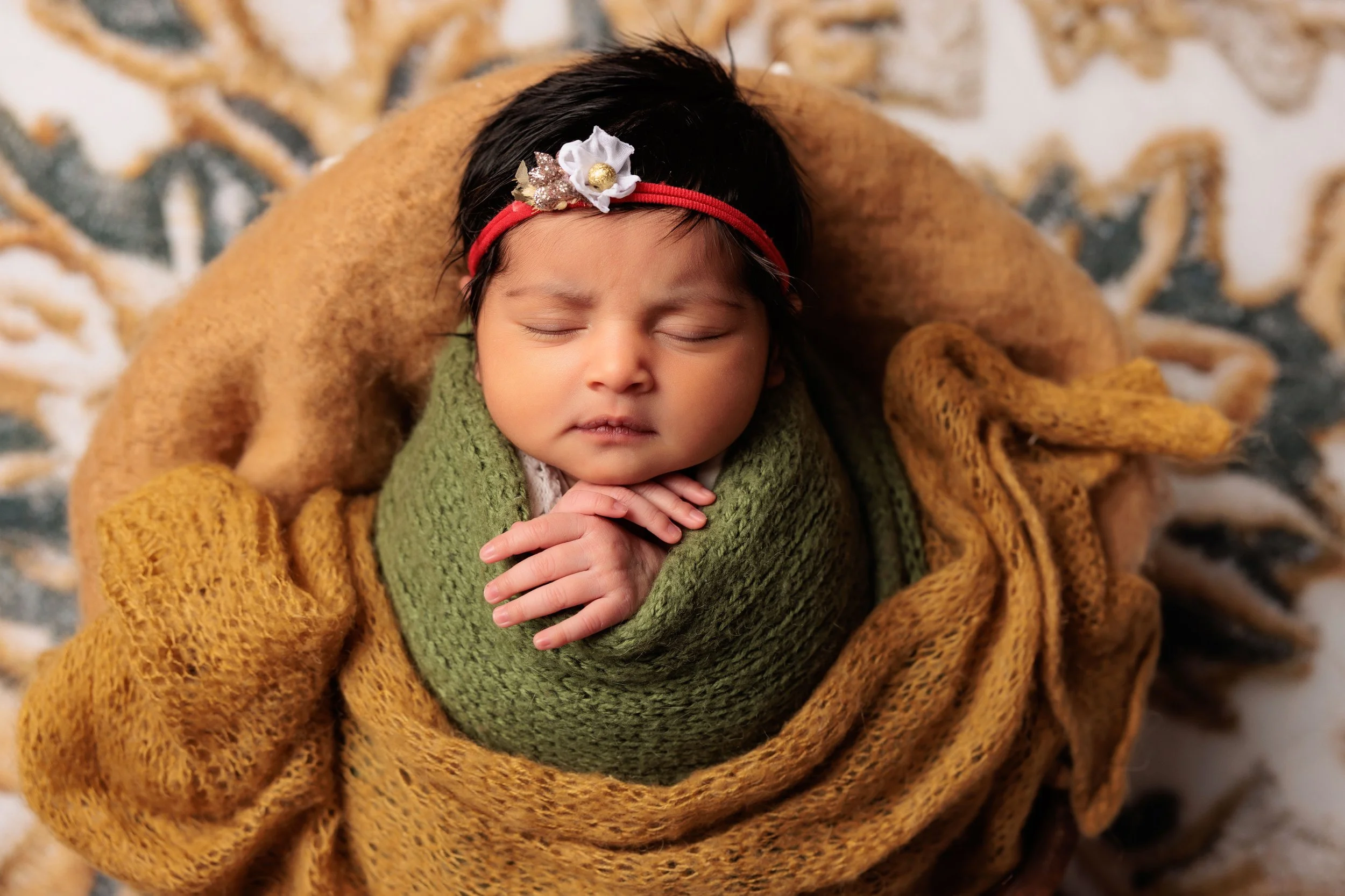 A sleeping baby girl with a red headband and a white flower, wrapped in green and brown knitted blankets, lying on a patterned surface.