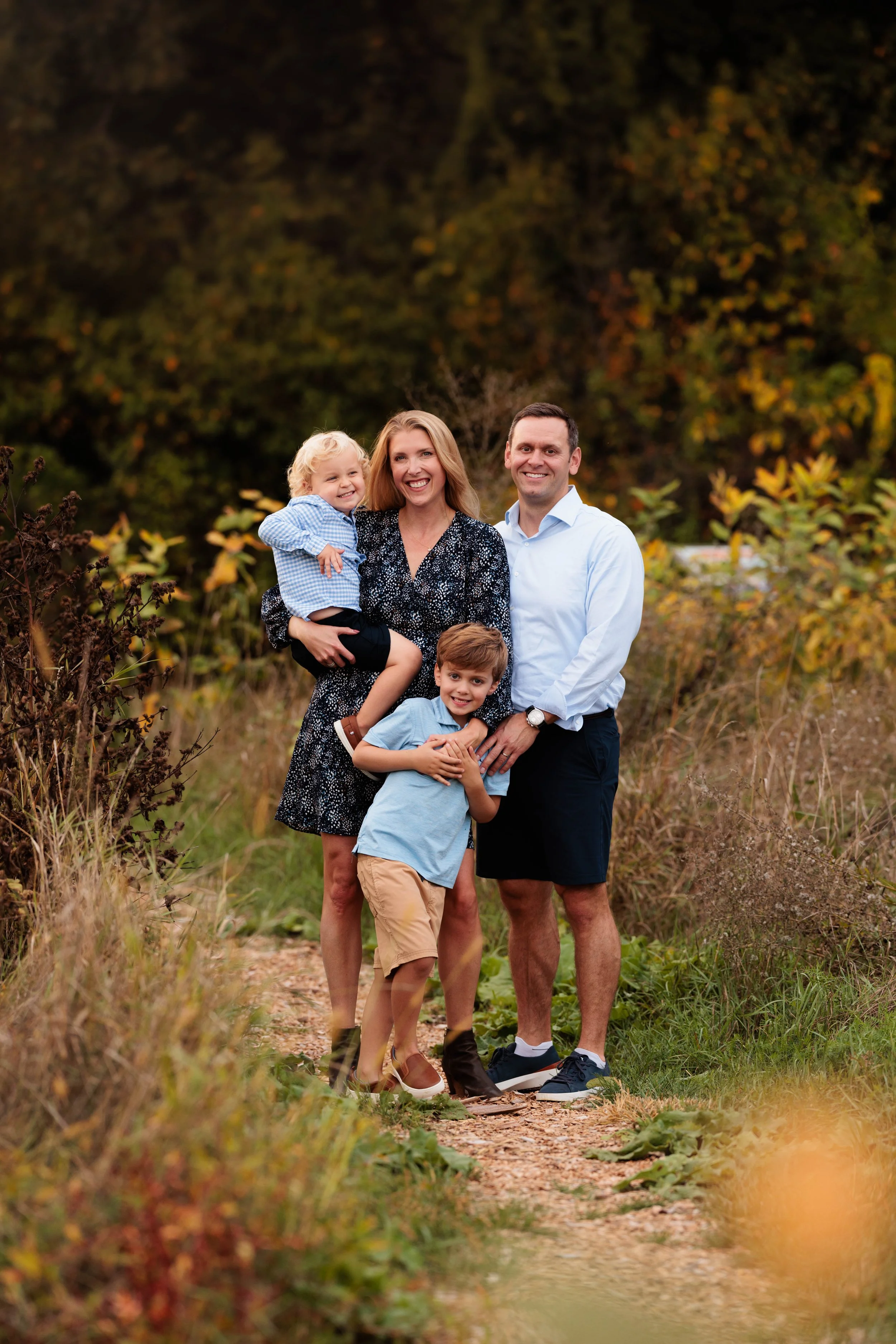Family of four standing on a trail outdoors during fall, smiling and posing for a photo. The family includes a woman, a man, a young boy, and a young girl.