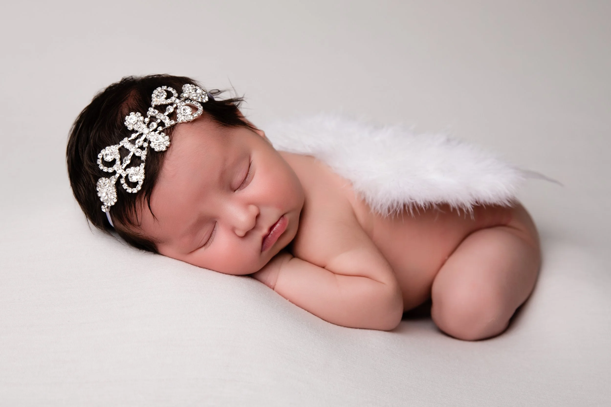 A peacefully sleeping newborn baby laying on a white surface, wearing a diamond-encrusted tiara and white feathered wings.
