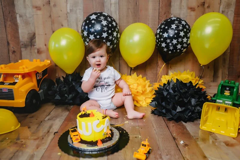 A young child sitting on a wooden floor during a birthday celebration with black and yellow balloons, paper decorations, and toy construction vehicles, including a cake with construction-themed decorations.