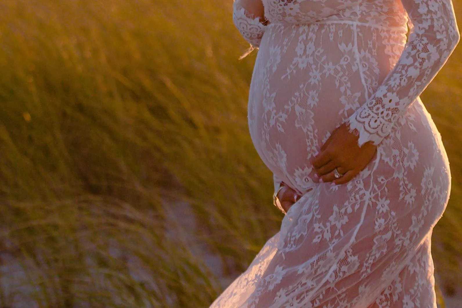 A pregnant woman wearing a white lace dress standing outdoors in a field during sunset.