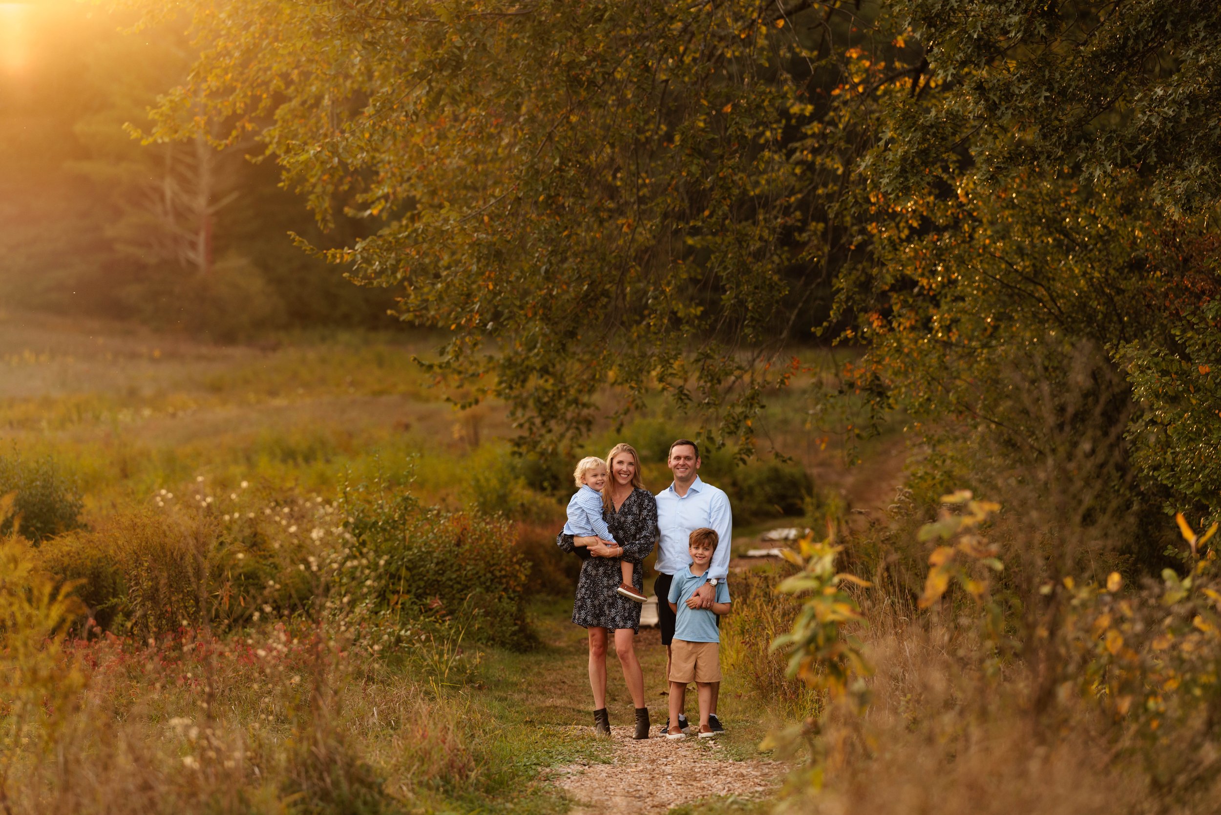 Family of four walking outdoors on a dirt trail surrounded by autumn foliage, with the sun setting in the background.