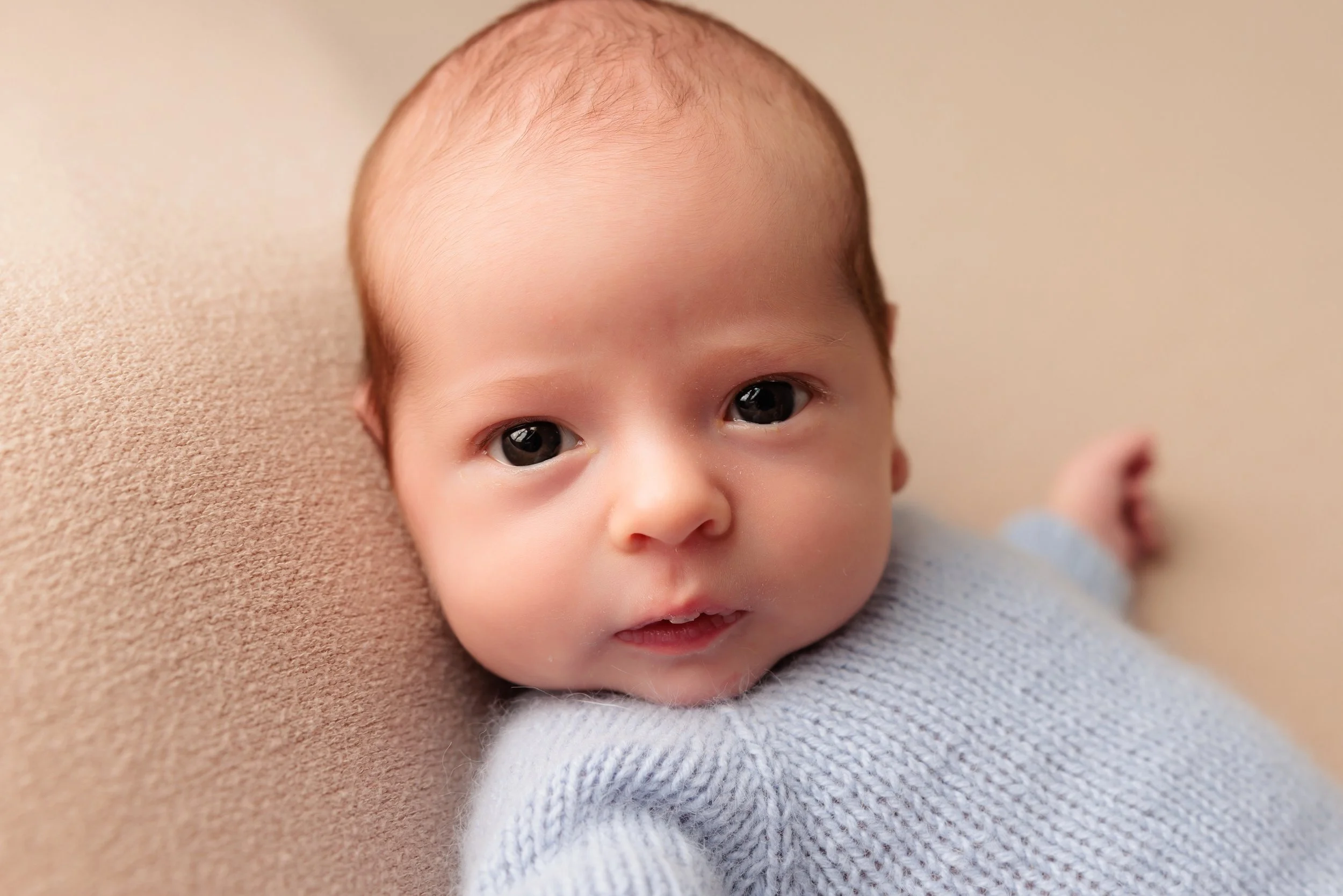 Close-up of a baby with brown hair and big dark eyes, lying on beige fabric, wearing a light gray sweater.