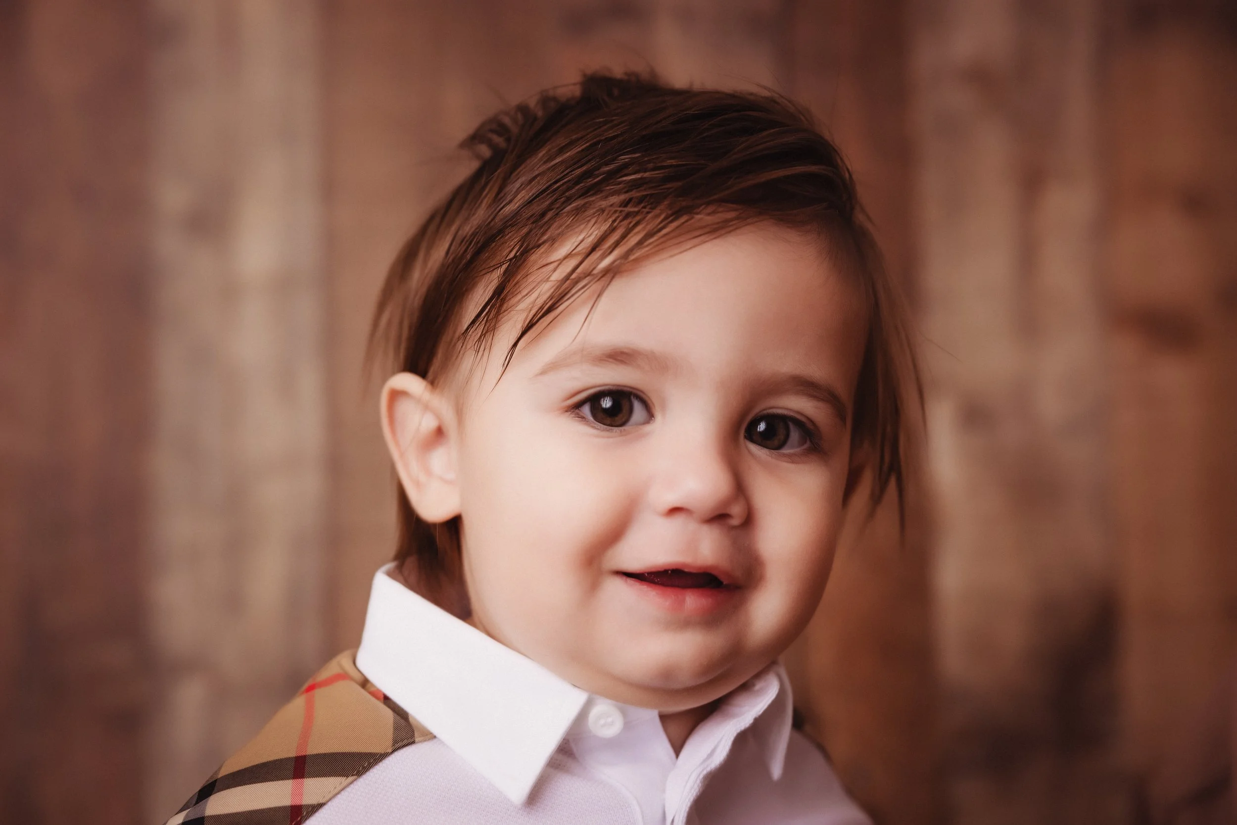 A young boy with wet brown hair wearing a white collared shirt with a plaid design on the shoulder, smiling and looking at the camera, with a wooden background.