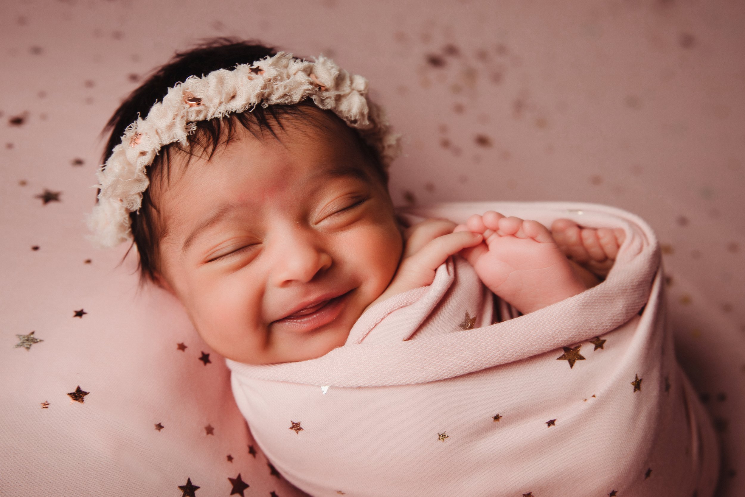 A smiling baby girl with closed eyes, wearing a floral headband, wrapped in a pink blanket with star patterns, lying on a matching star-patterned blanket.