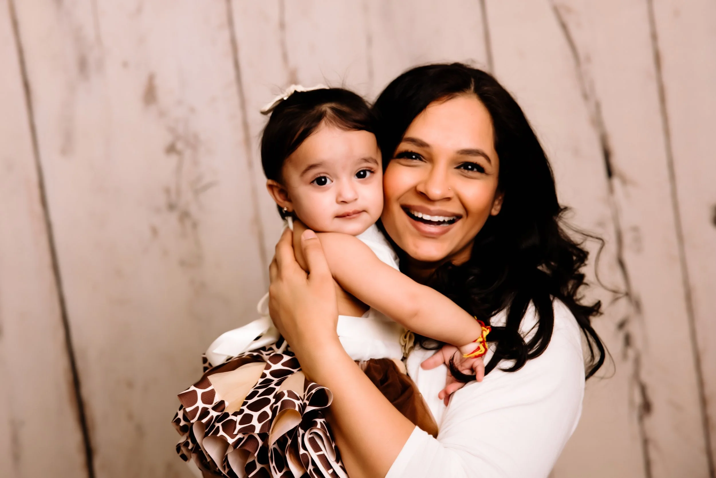 A woman with dark hair holding a young girl with dark hair and a white bow, both smiling and embracing each other, against a rustic wooden background.