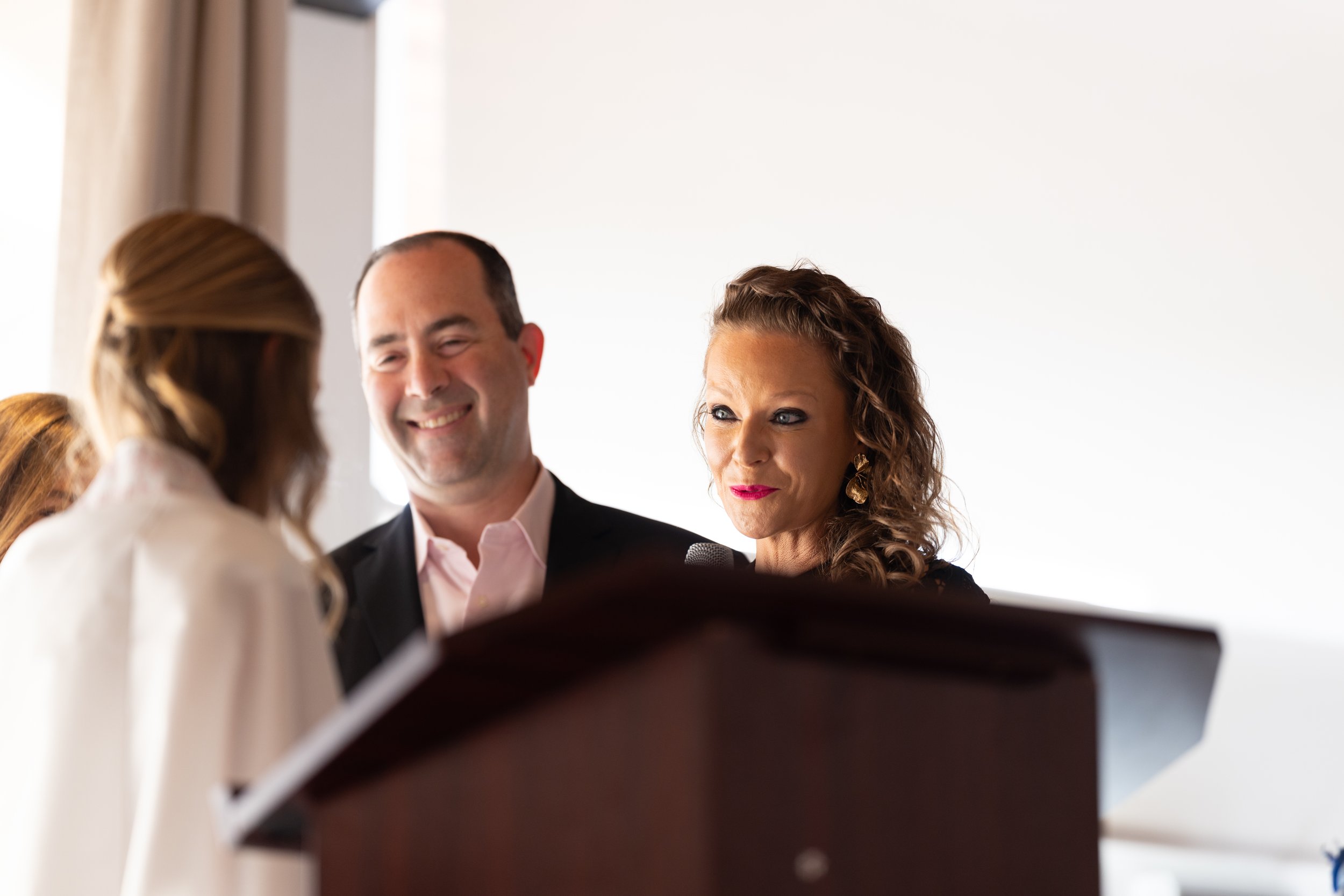 A woman and man standing next to each other, smiling and looking at a woman with her back to the camera, in a bright room.