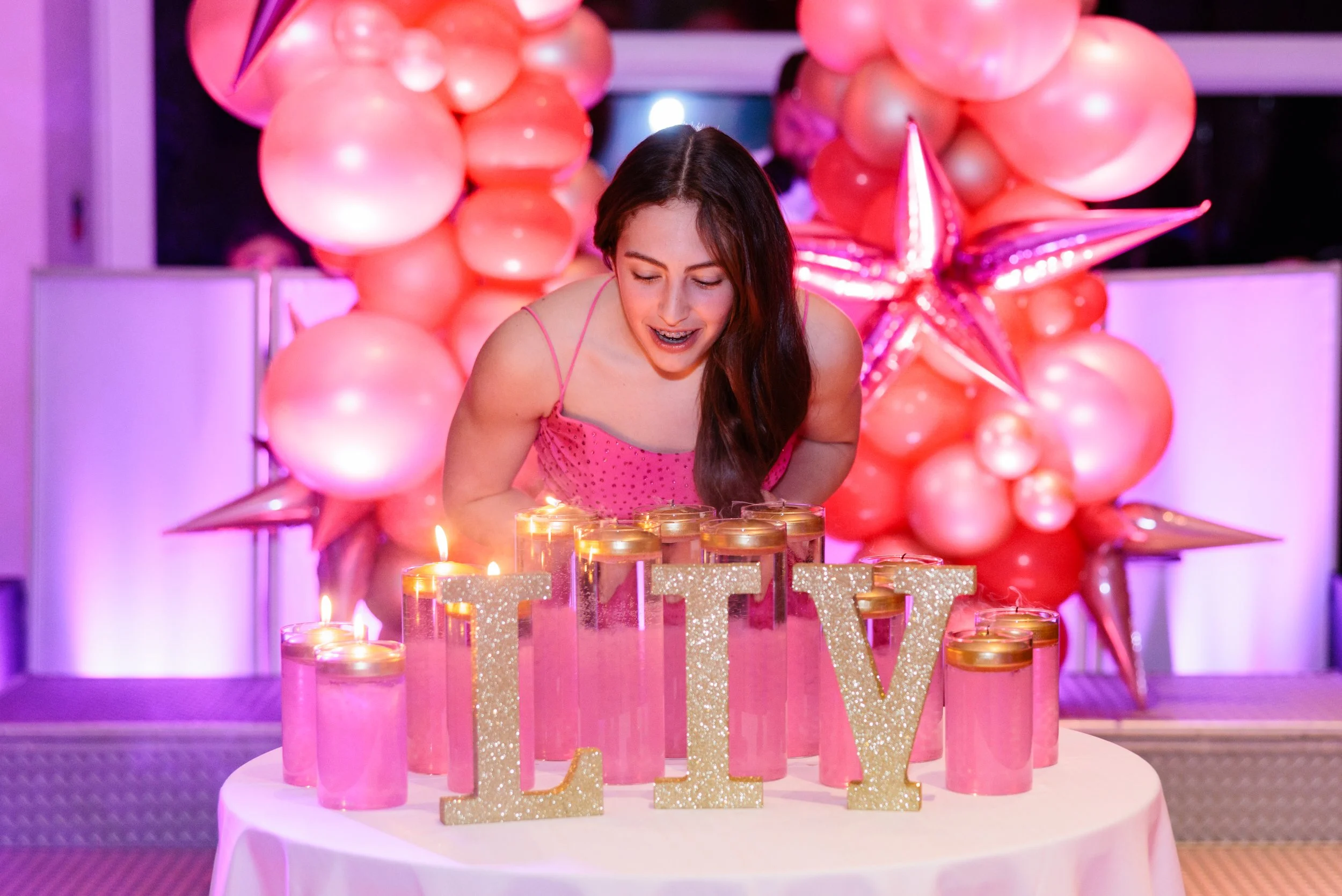 Young woman in pink dress blowing out candles on pink birthday cake with candles spelling 'LOVE' amidst pink balloons and star decorations.