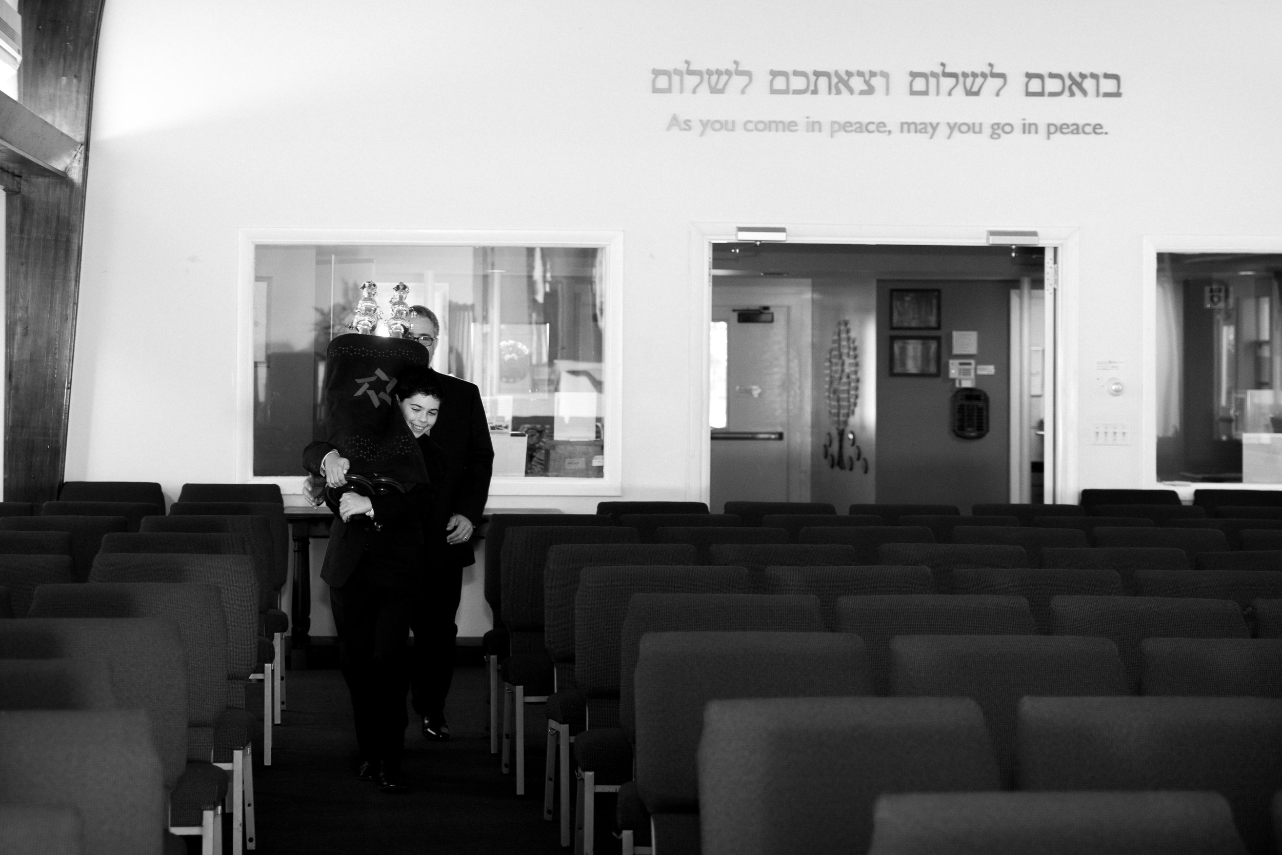 A woman and girl walking down an aisle in an empty conference or event room with chairs, floral decorations, and a window with a sign in Hebrew and English that says, 'As you come in peace, may you go in peace.'