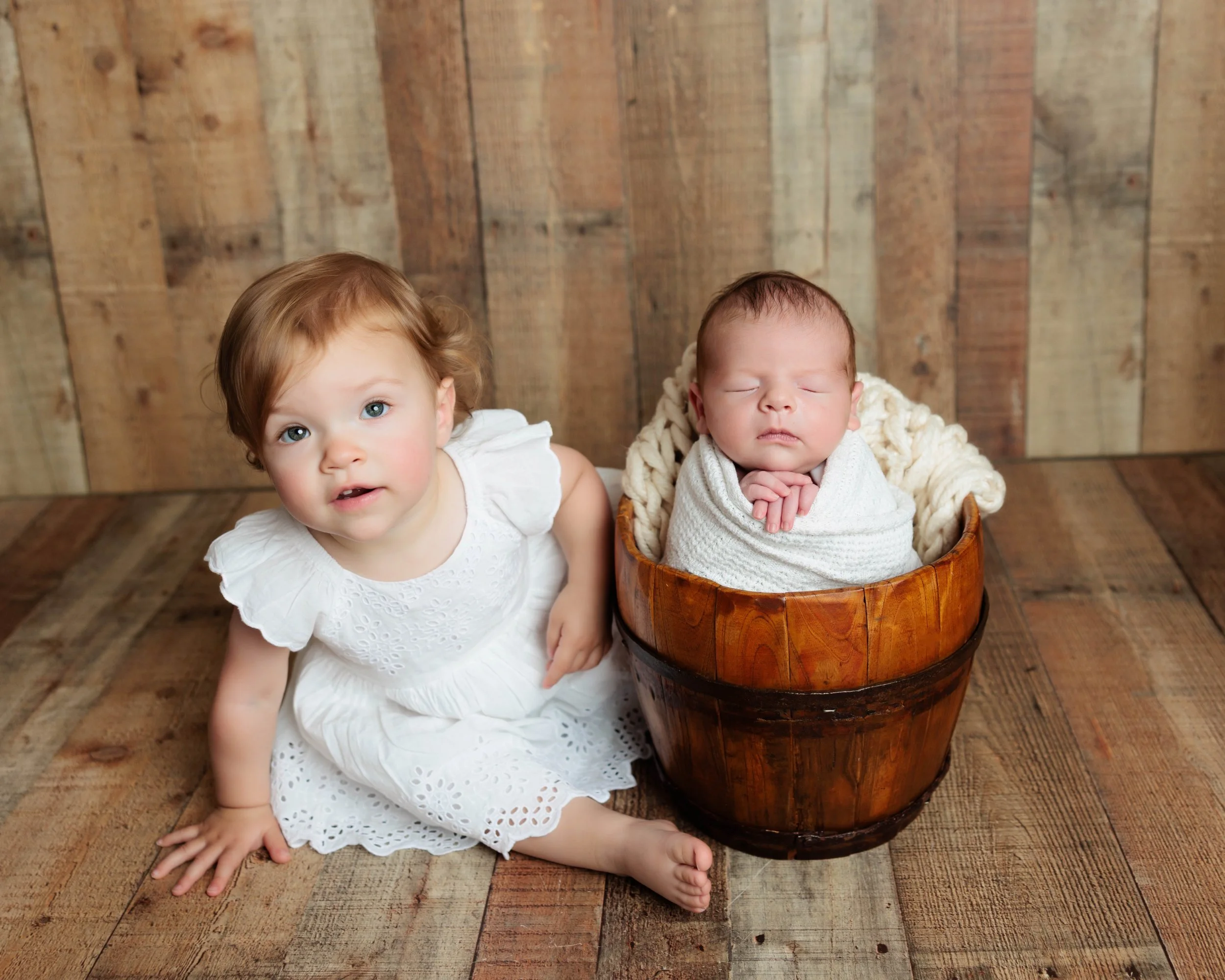 A young girl with brown hair and blue eyes sitting on a wooden floor next to a wooden bucket with a sleeping newborn wrapped in a blanket inside.