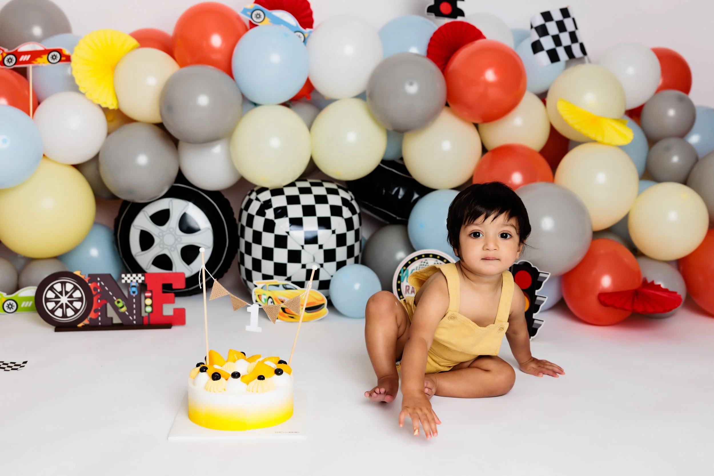 A young child in a yellow outfit sitting on the floor in front of a colorful balloon backdrop and a cake decorated with yellow and black accents, with birthday-themed decorations around.