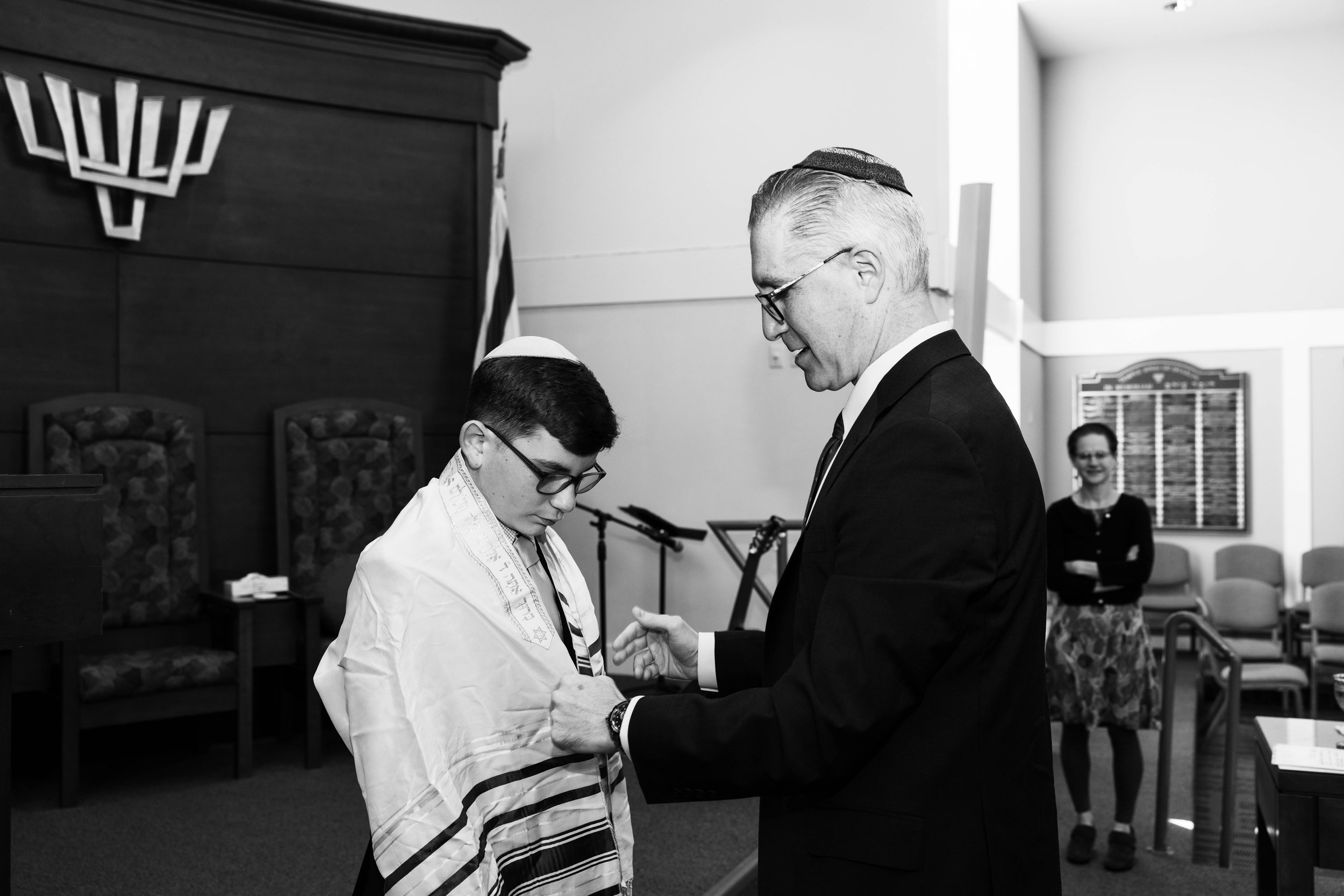 A young boy wearing a tallit and kippah receives a blessing from an older man in a suit during a Jewish religious ceremony, with a woman watching and smiling in the background.