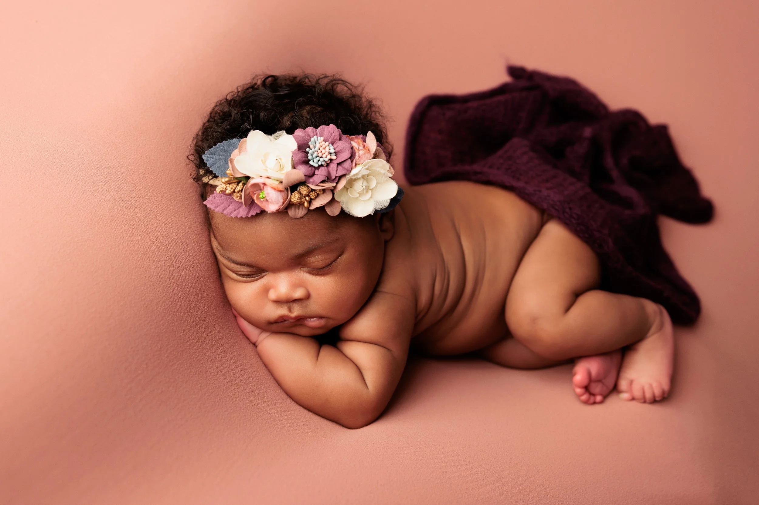 A sleeping baby with a floral headband lying on a pink surface, partially covered with a dark purple cloth.