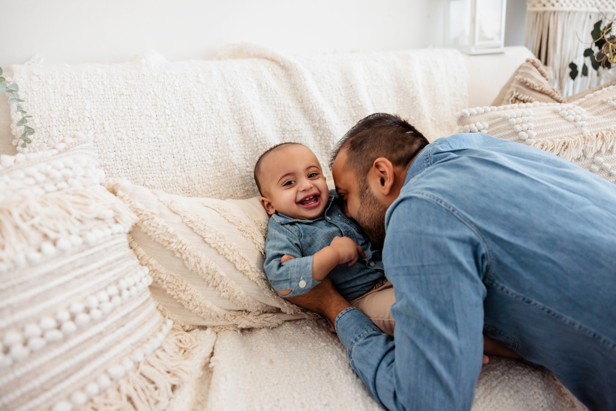 A man and a young child lying on a cream-colored sofa, playing and smiling at each other.