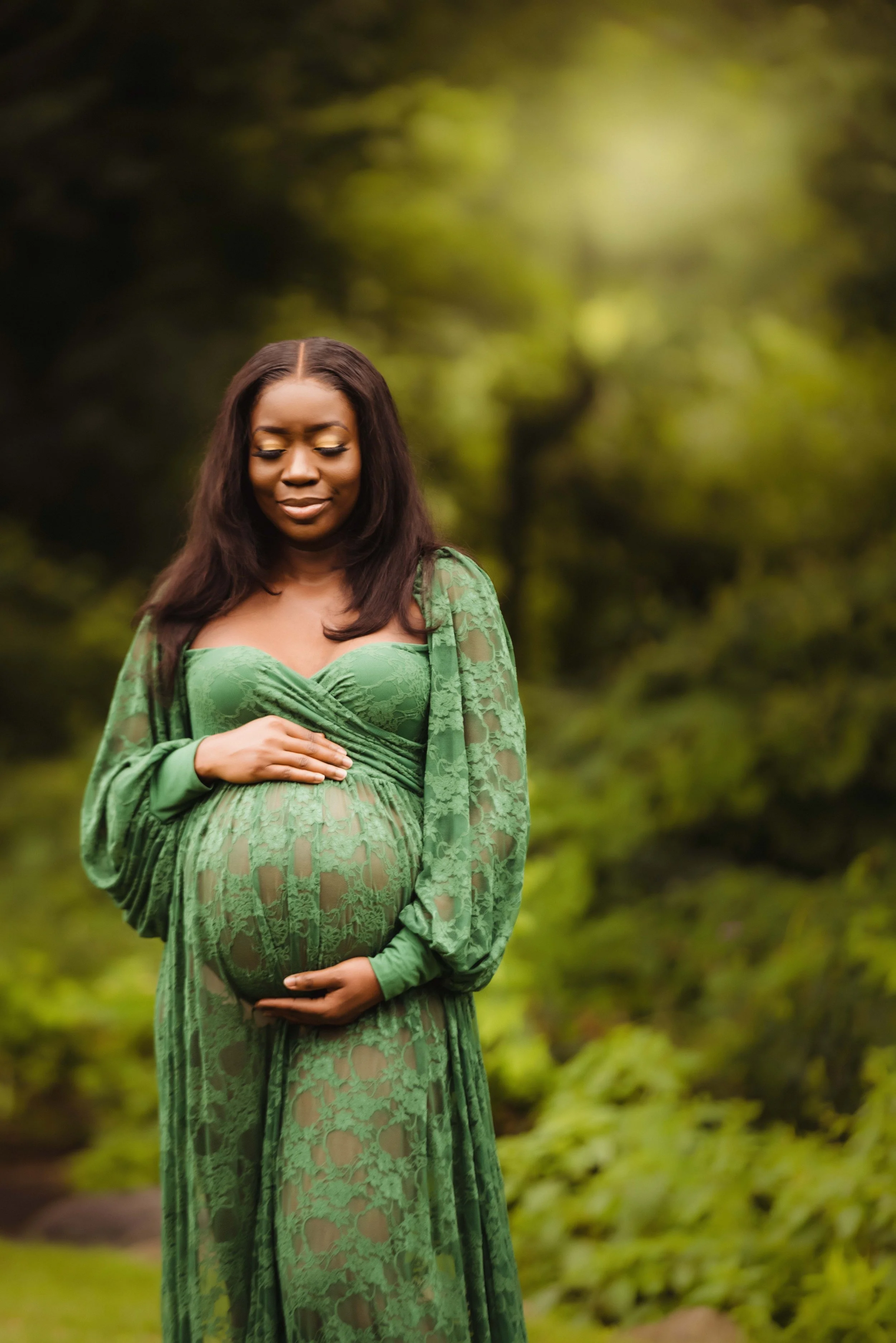 A pregnant woman in a green lace dress standing outdoors in a natural setting, gently holding her belly with her eyes closed.