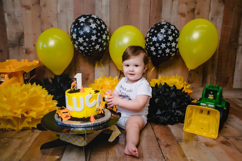A young child sitting on a wooden floor celebrating their first birthday with a yellow and black construction-themed cake, surrounded by yellow and black balloons, paper decorations, and toy construction vehicles.