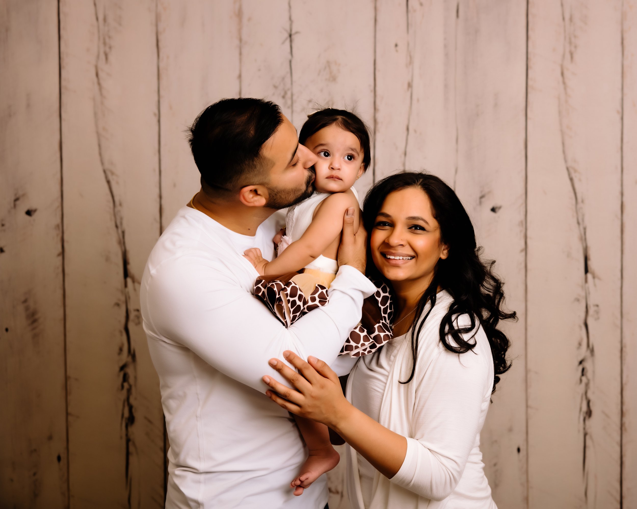 A family of three with a father, mother, and young daughter, all smiling and hugging against a wooden background.