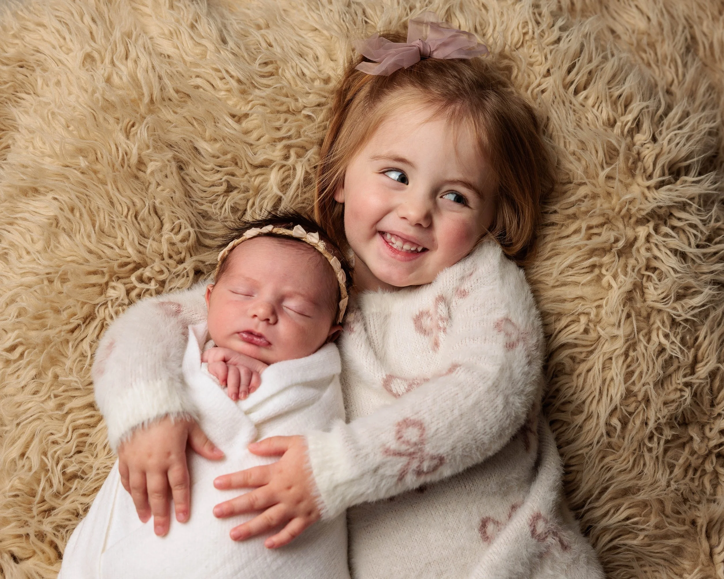 A young girl with a big pink bow in her hair hugging a sleeping baby dressed in white, lying on a fluffy beige rug.