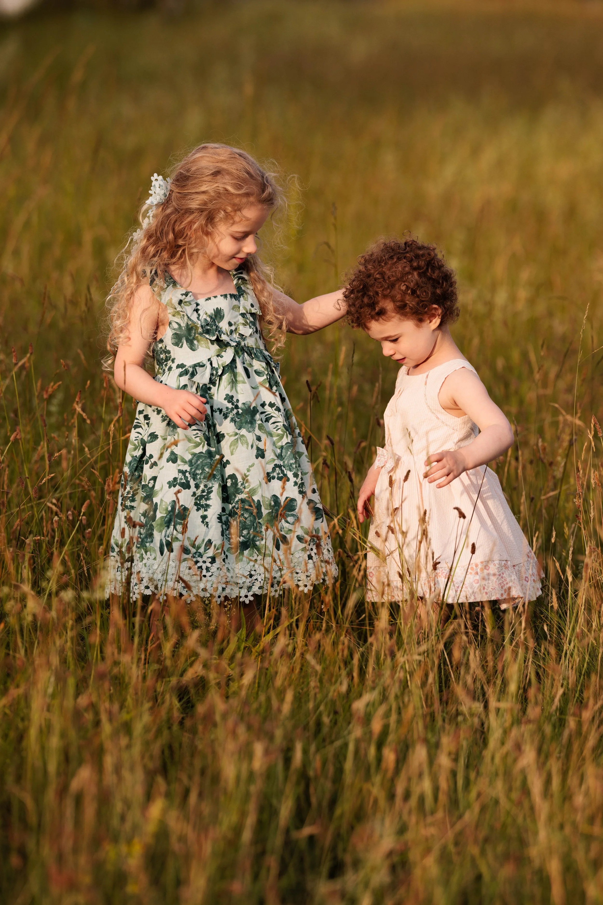 Two young girls with curly hair, one blonde and one brunette, standing in a grassy field during golden hour, wearing light summer dresses and engaging with each other.