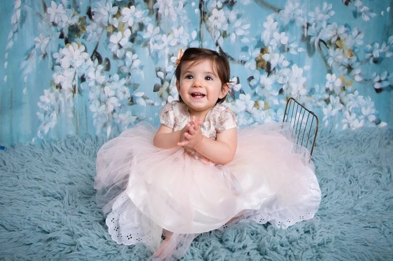 A young girl in a pink tulle dress sitting on a fluffy blue rug, smiling with hands clasped, in front of a blue floral backdrop.