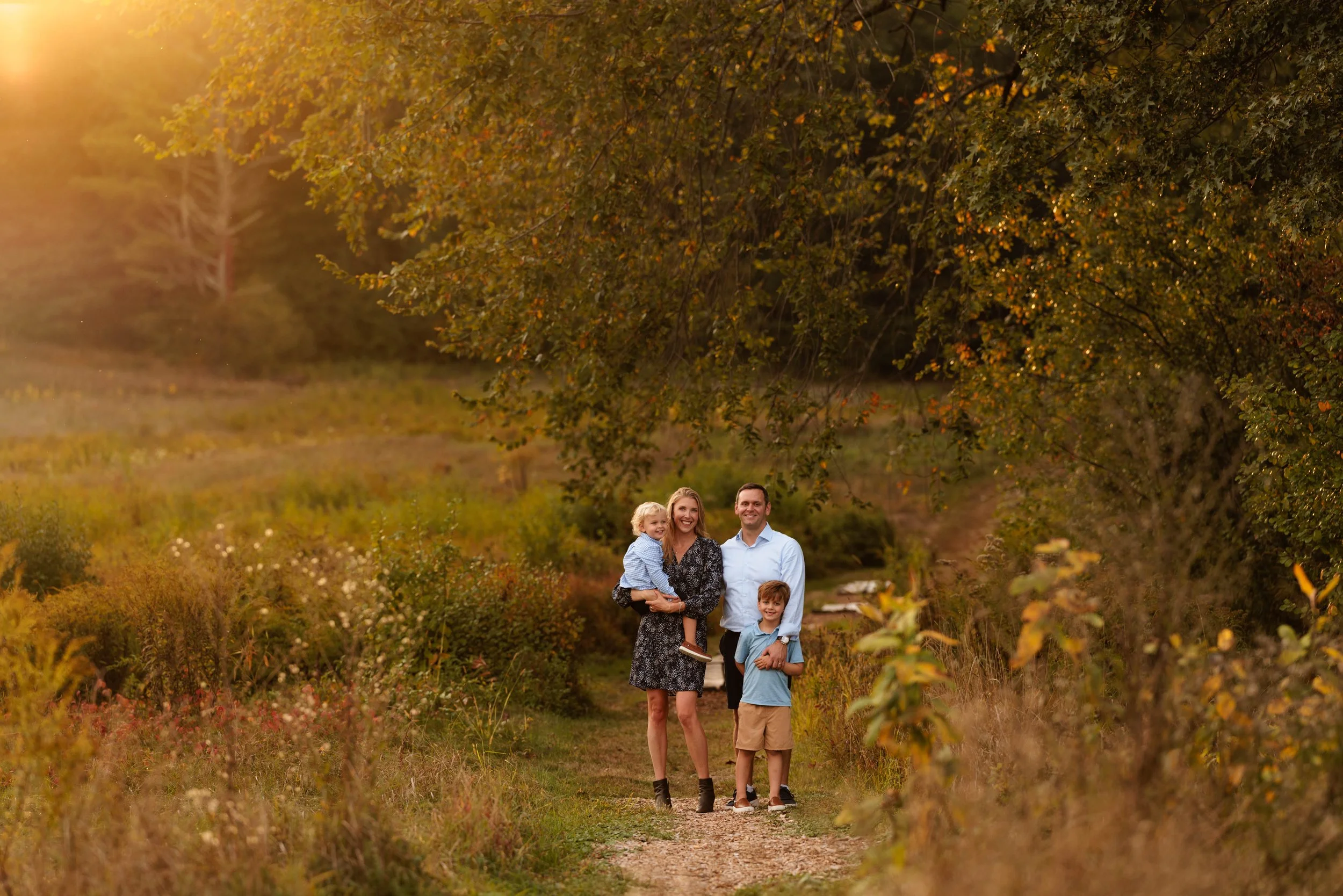 A family of four, including two children, walking on a dirt trail through a natural landscape with autumn leaves, trees, and sunlight filtering through the branches.