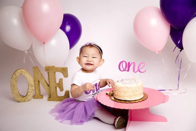 A happy baby girl celebrating her first birthday, sitting on the floor with a cake and pink, purple, and white balloons around her, with "ONE" glittery letters and a pink "one" cake topper.