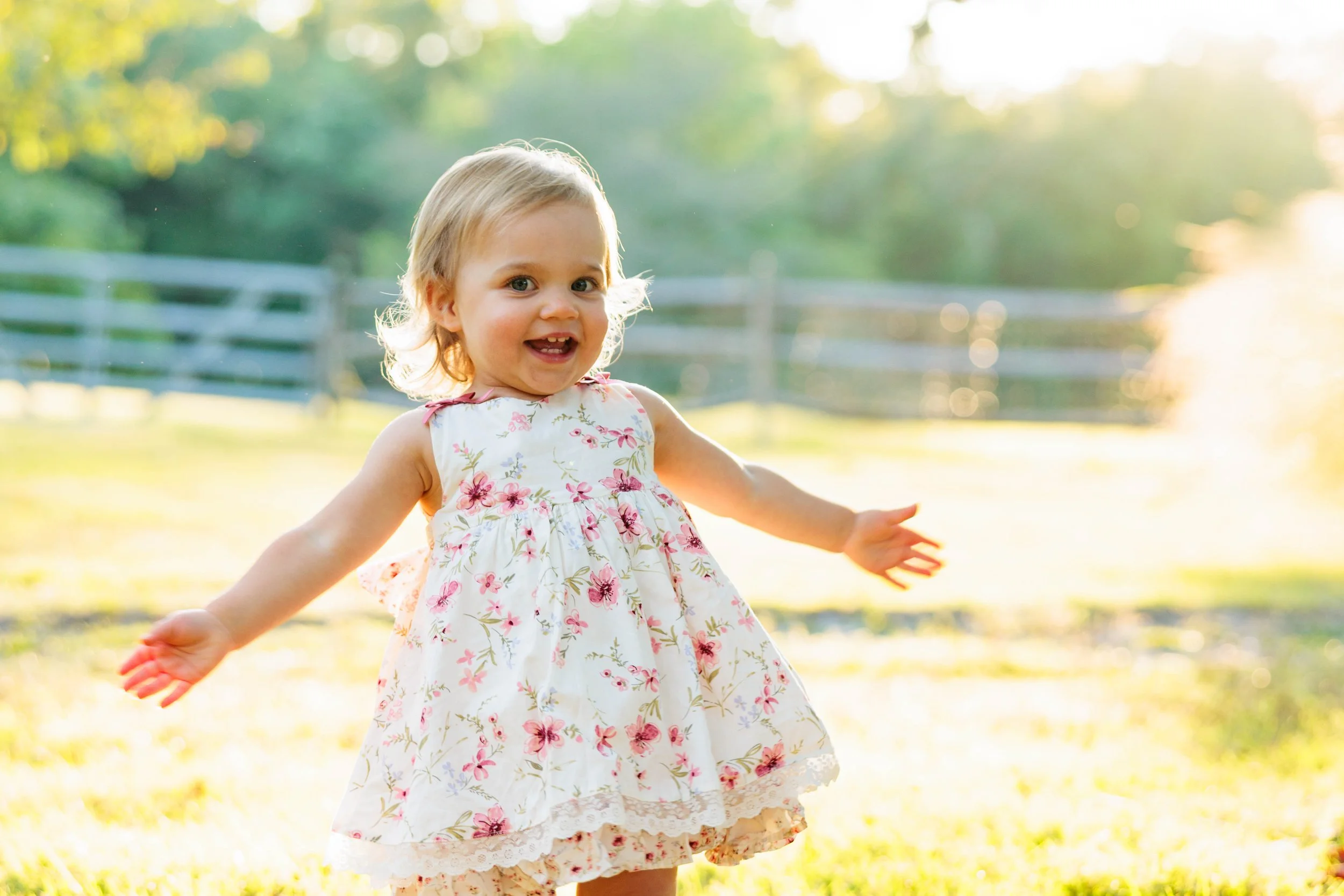 A young girl in a floral dress playing outside, smiling with her arms outstretched in a sunny, grassy field.