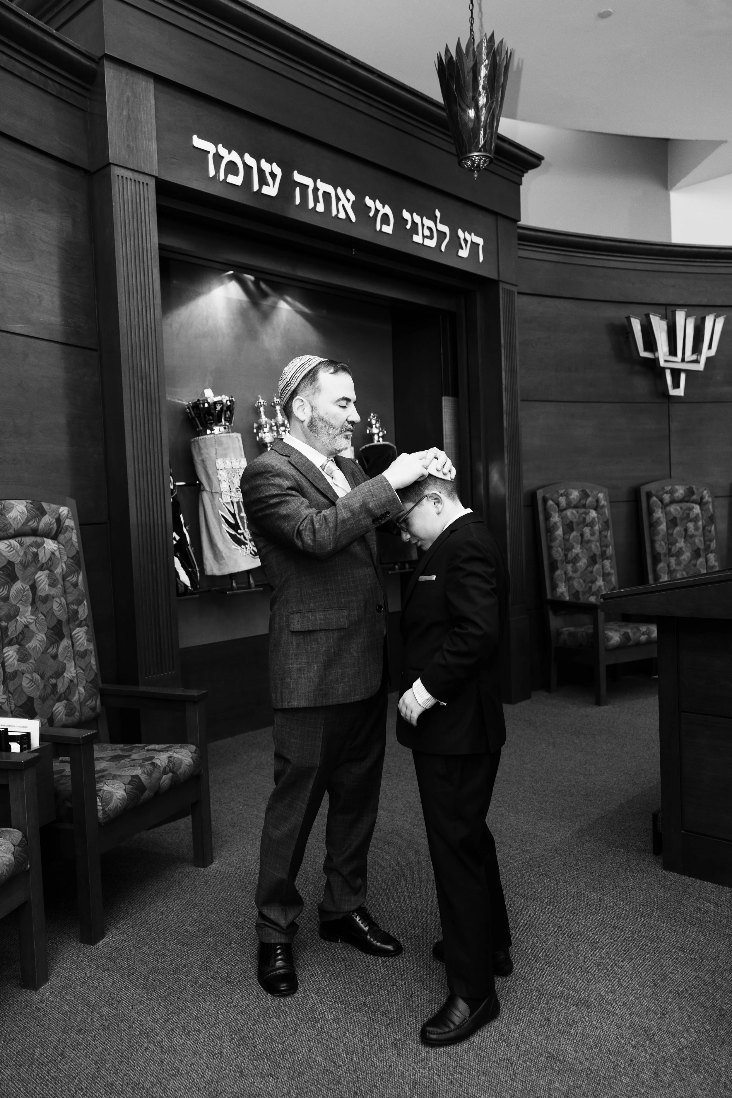 A Jewish man in a suit and kippah blessing a boy in a suit and glasses with a hand on his head in a synagogue, with Hebrew text on the wall behind them.