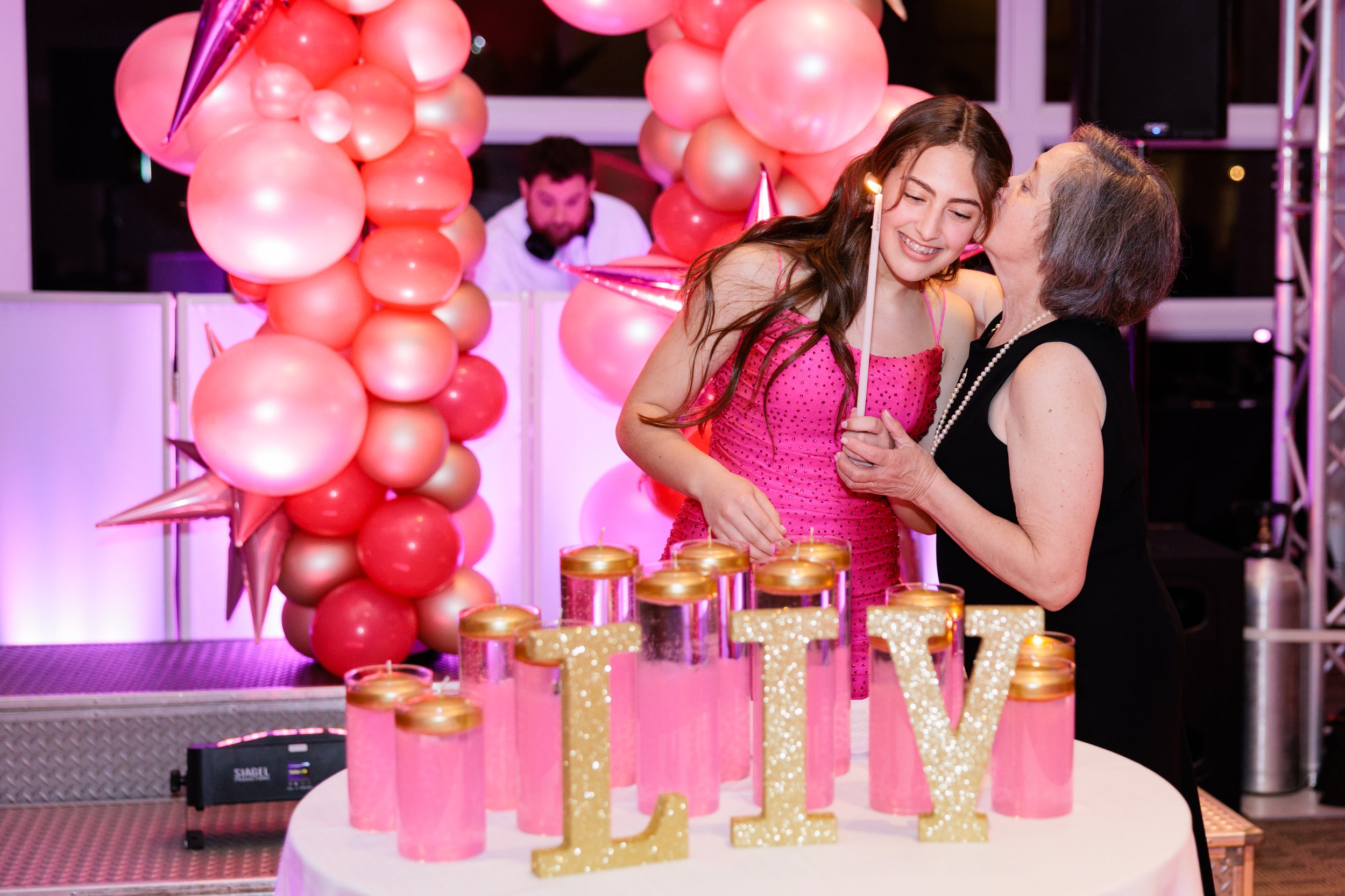 A young woman in a pink dress and an older woman in a black dress sharing a kiss at a birthday celebration with pink balloons, gold candles, and gold glittered word 'LIV' on a cake table.