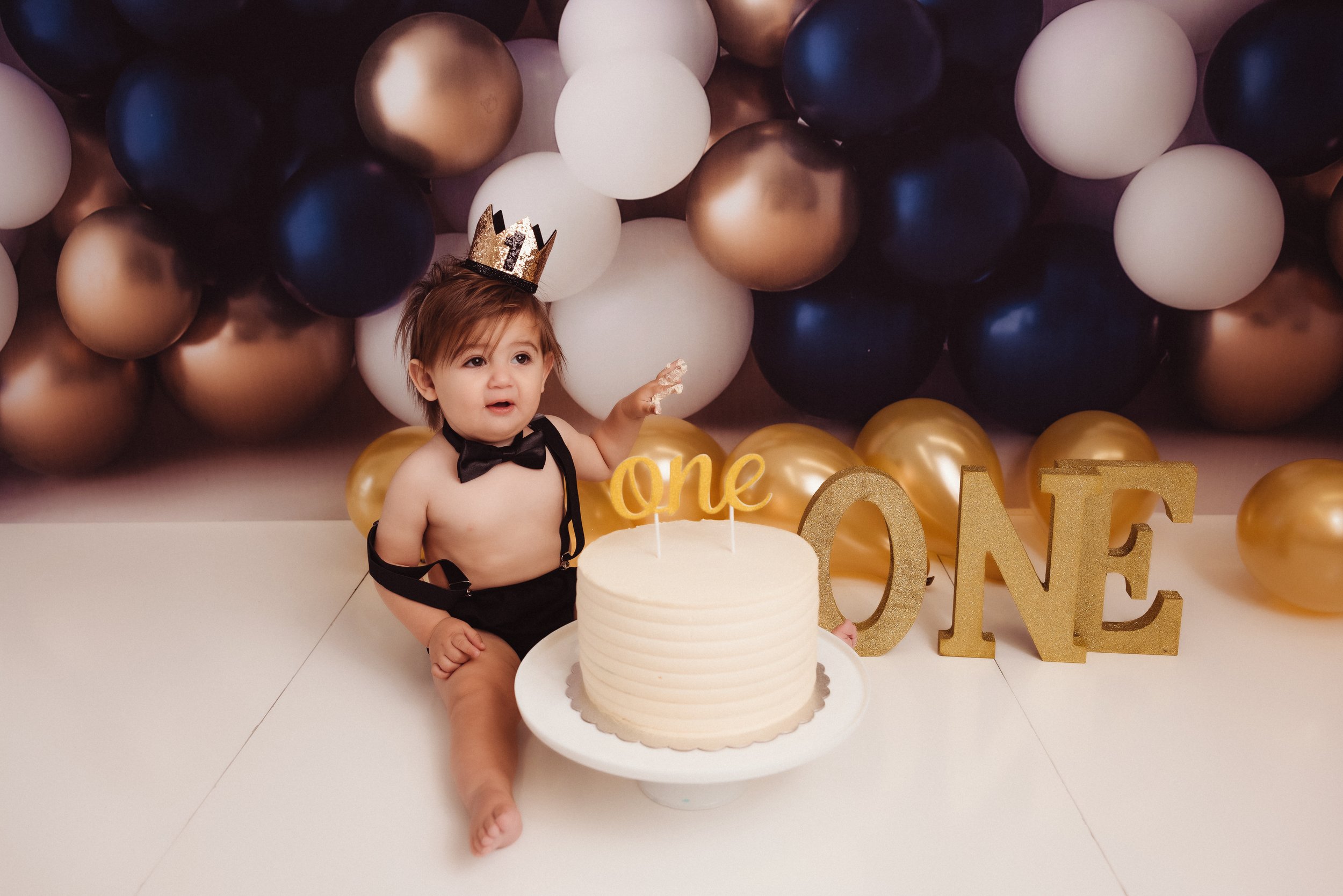 A toddler celebrating their first birthday, sitting on the floor in front of a white birthday cake with three candles, surrounded by gold, white, black, and copper balloons, with a "one" and "one" sign, wearing a black bow tie and suspenders, with a 