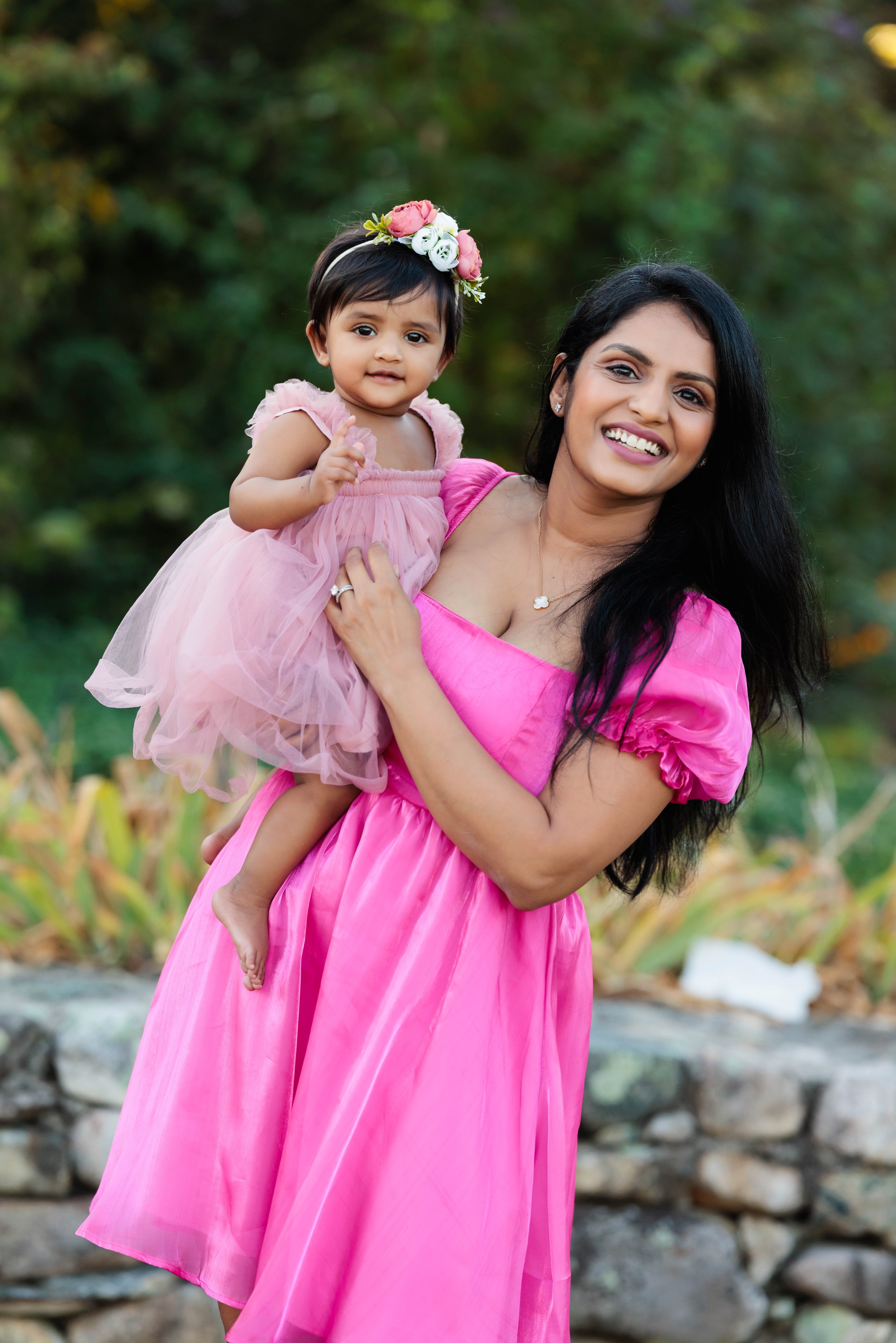 Smiling woman in a pink dress holding a young girl in a matching pink dress with a flower crown in an outdoor setting.
