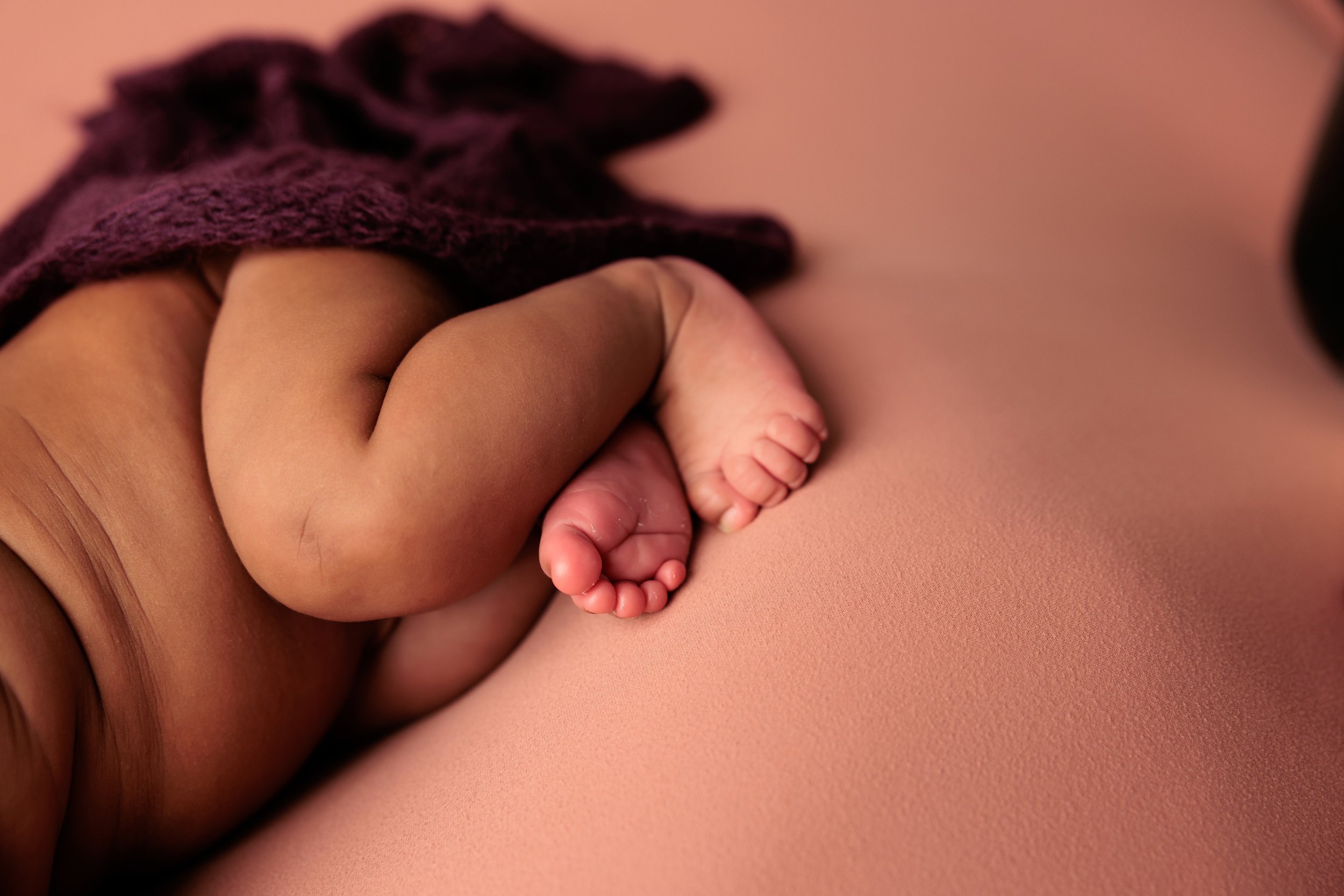 Close-up of a newborn baby's tiny feet and hand, lying on a pink surface, partly covered with a purple cloth.