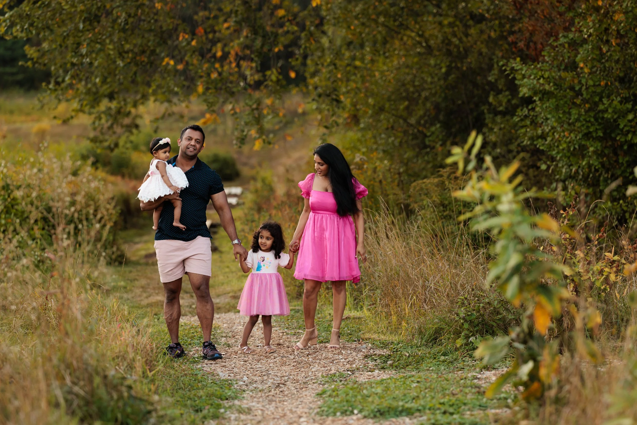 Family walking on a nature trail surrounded by trees and grass, with the father carrying a young girl in a white dress, the mother in a pink dress, and an older girl holding her parents' hands.