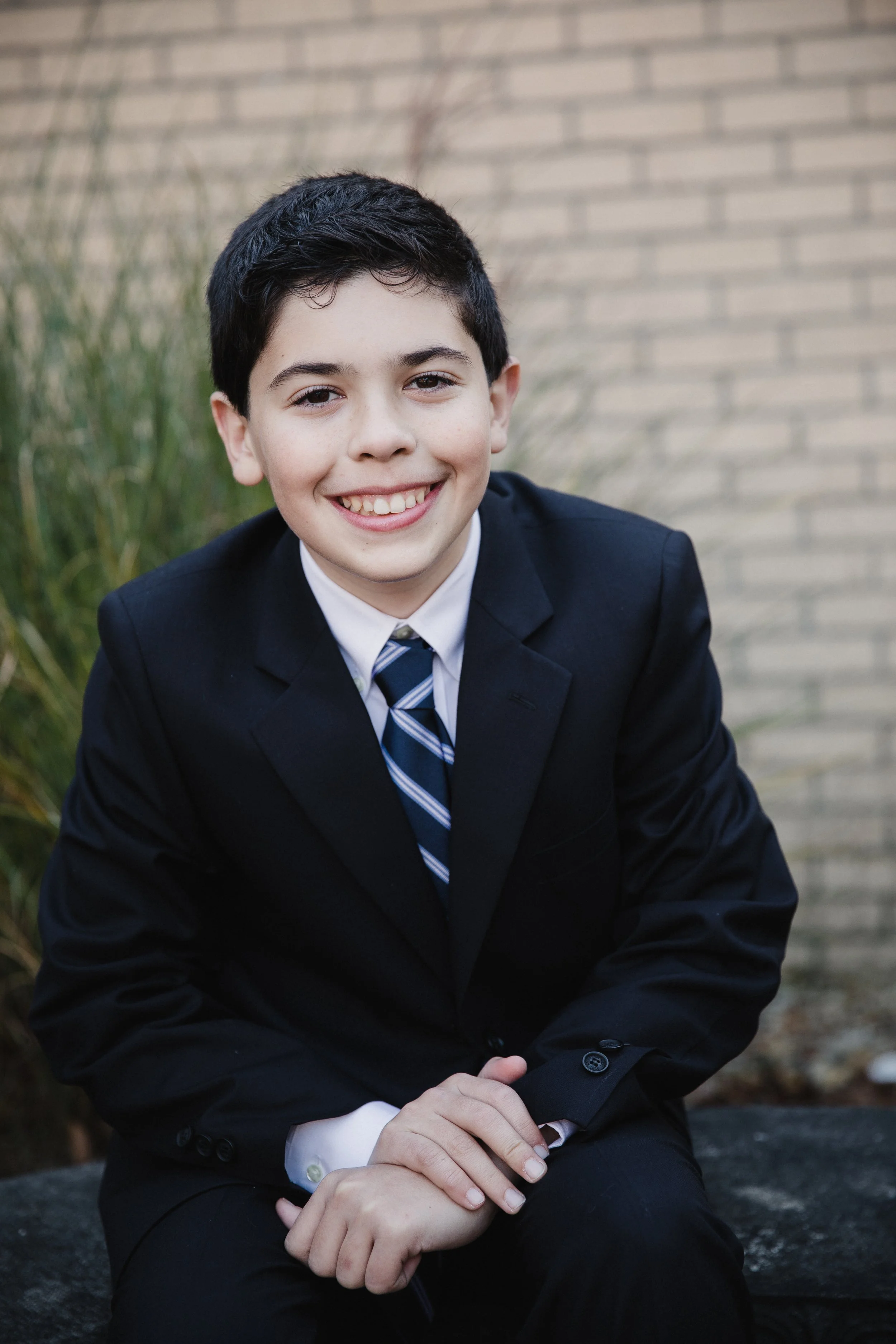 A young boy in a navy blue suit and striped tie smiling while sitting outdoors against a brick wall and greenery.