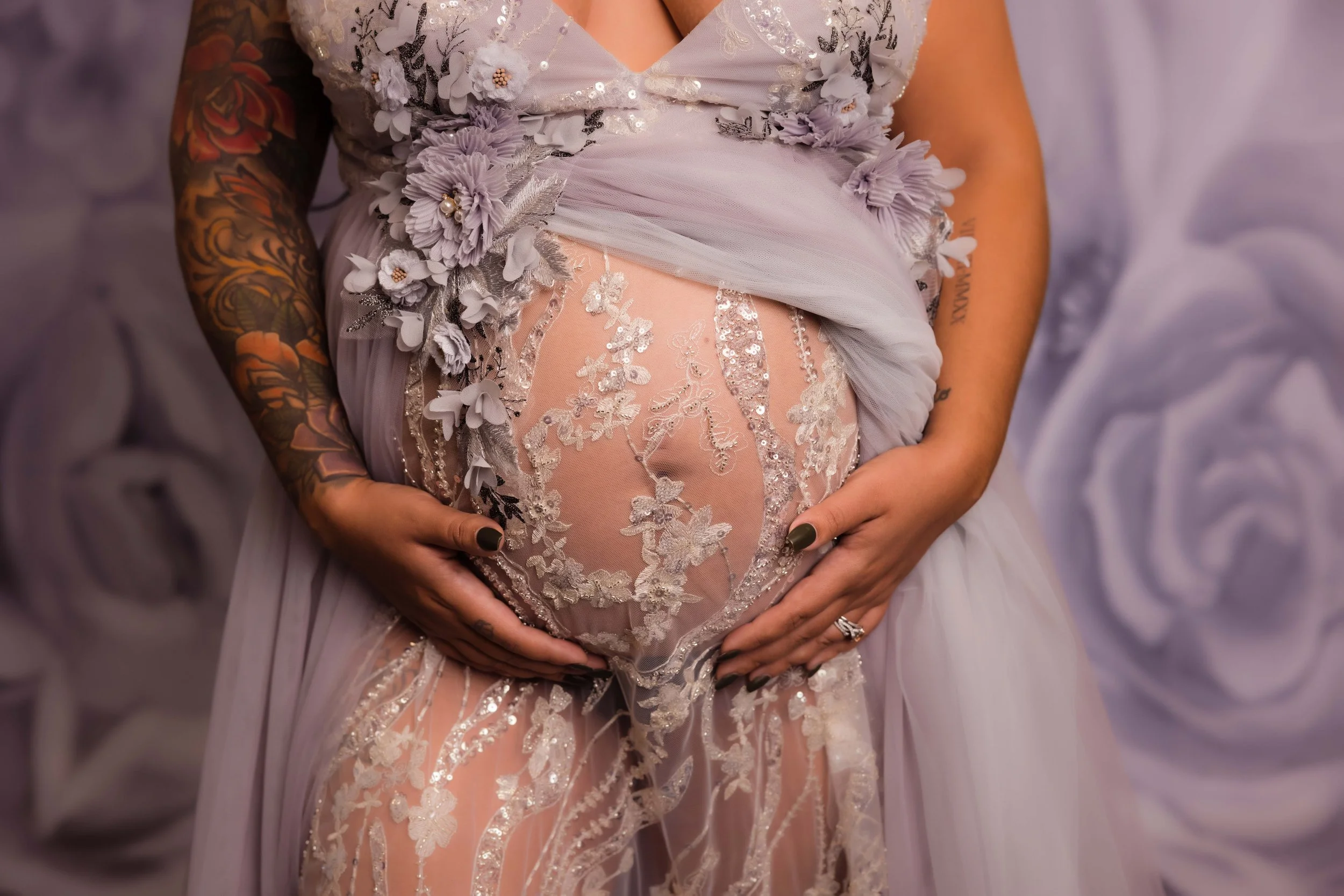 Close-up of a pregnant woman in a lavender dress with lace and floral embellishments, holding her belly with both hands, revealing tattoos on her arms and a ring on her finger, in front of a lavender floral backdrop.