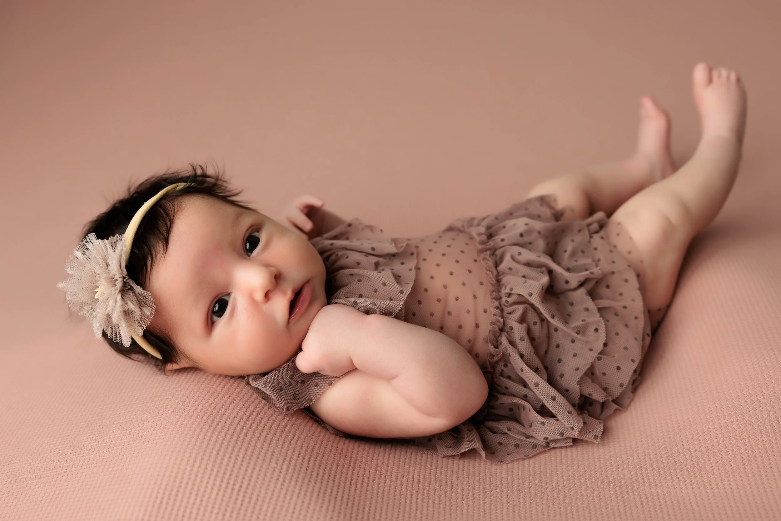 A baby girl lying on a pink textured surface, wearing a beige polka dot dress and a flower headband, looking at the camera with one hand near her face.