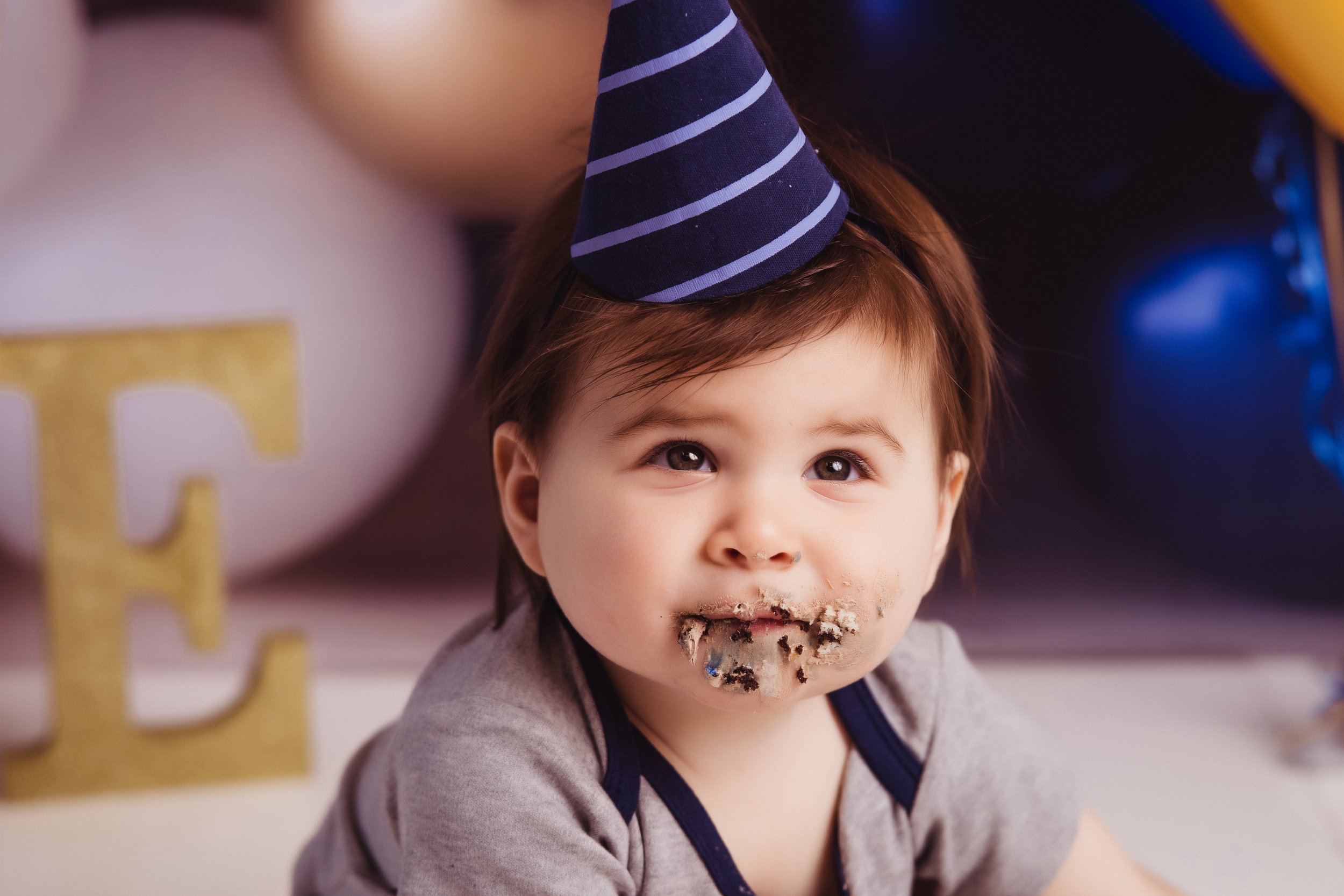 Young child with birthday cake and crumbs on face, wearing a blue striped party hat.